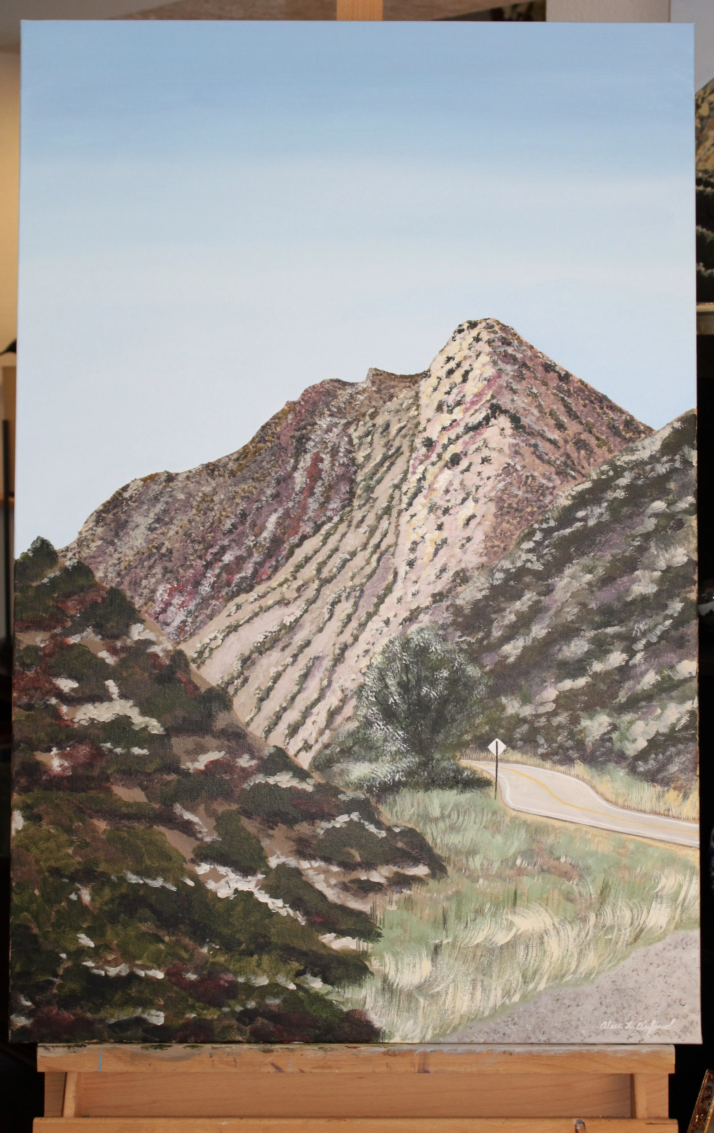 Alice Oxford's process photo of a painting of a mountainous landscape with a winding road, green vegetation, and a clear blue sky.