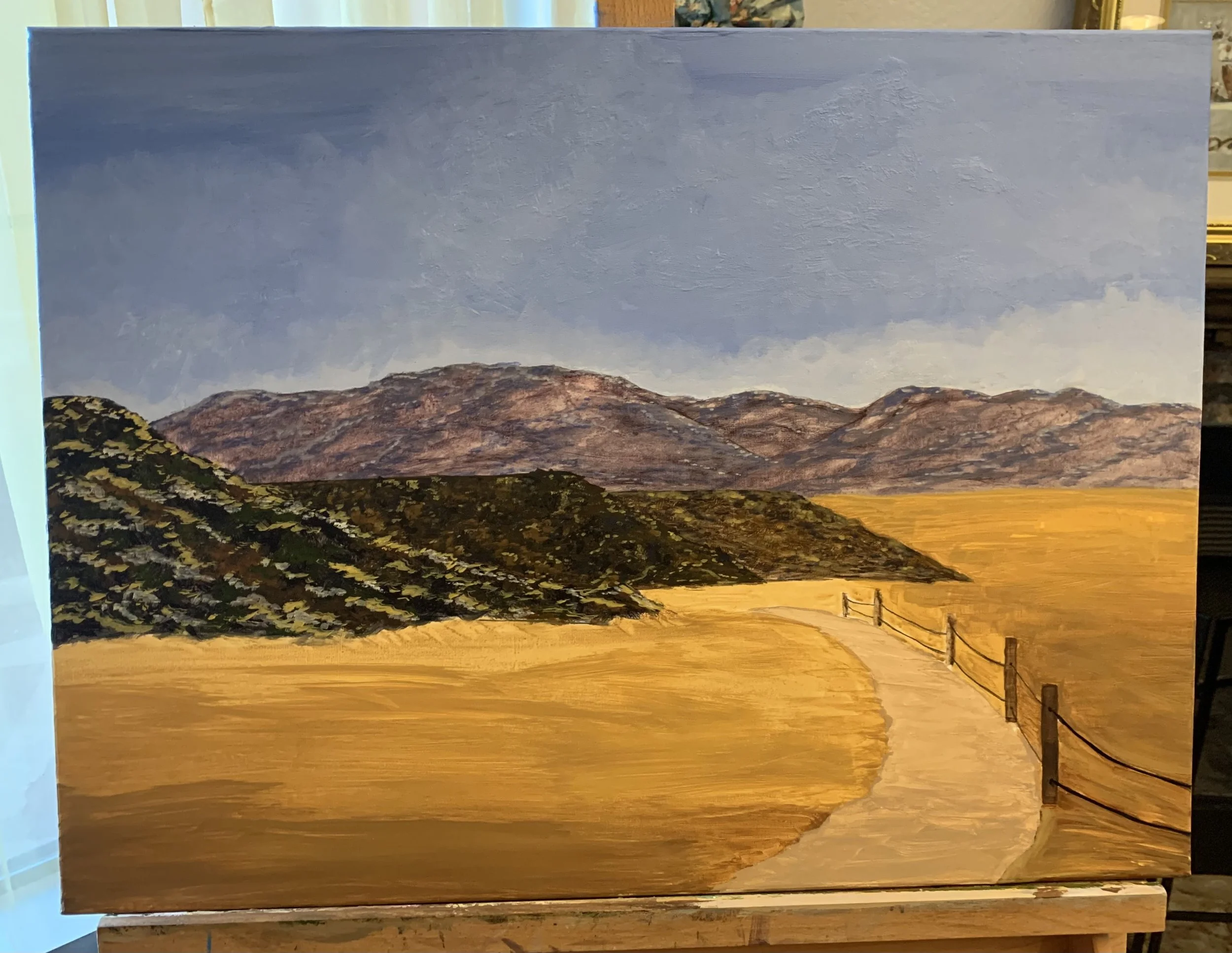 Alice Oxford's process photo of painting of a natural landscape featuring a winding path leading through a field with hills in the background, under a cloudy sky.