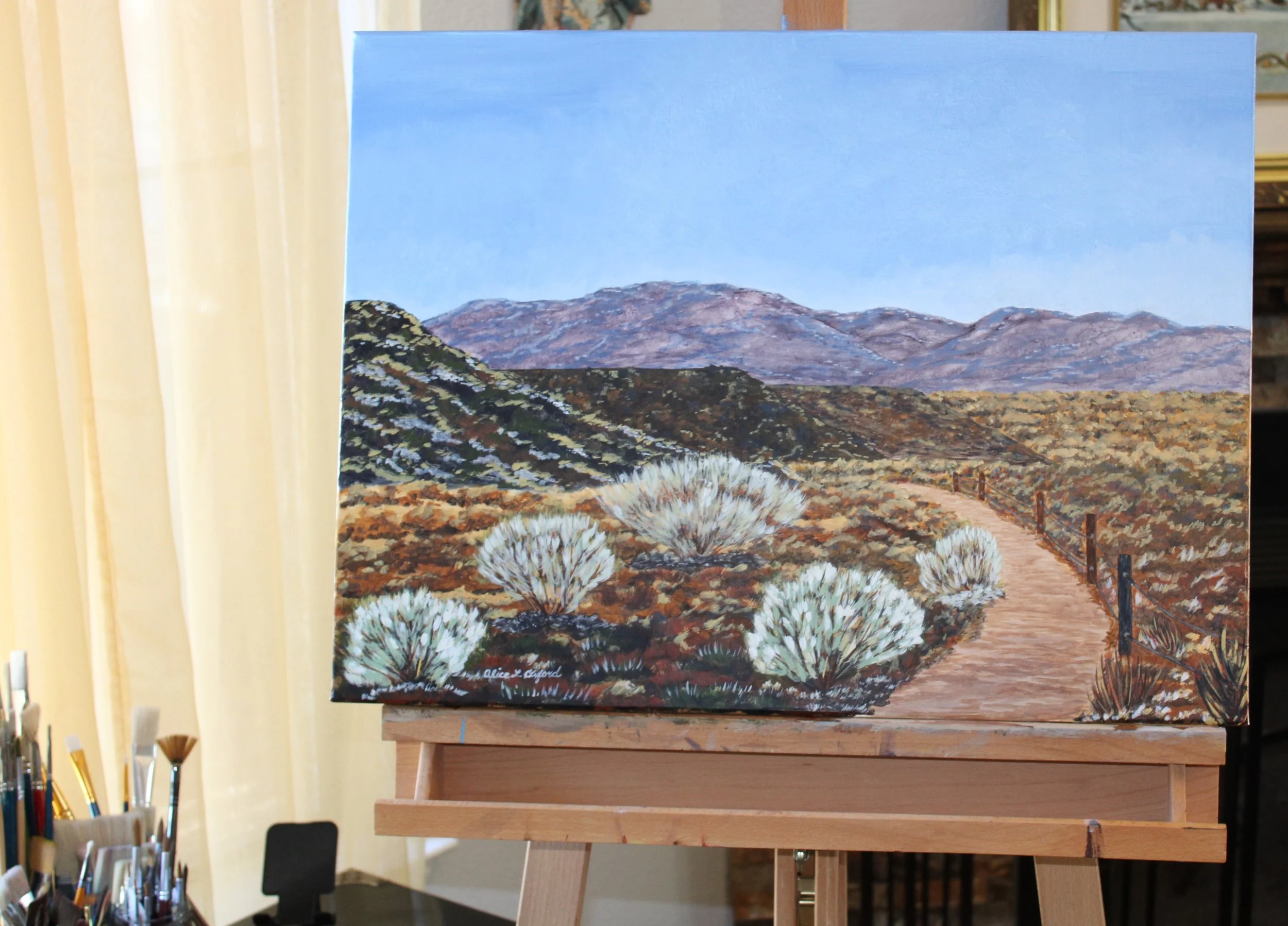 Alice Oxford's process photo of landscape painting on easel depicting desert scenery with a dirt path, shrubs, and distant mountains under a blue sky.
