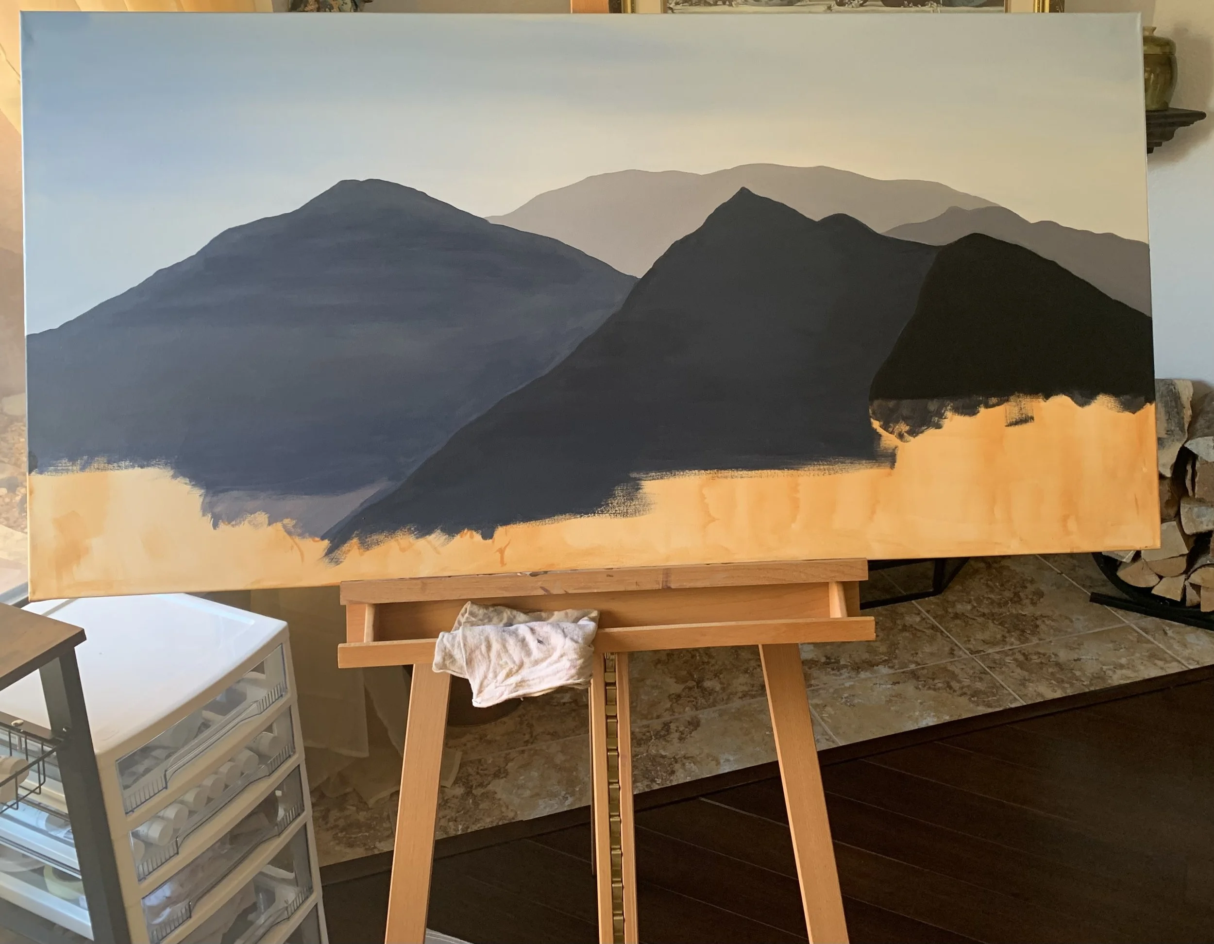 Alice Oxford's process photo of unfinished landscape painting on an easel, featuring dark mountains against a light sky, with some areas not fully painted.