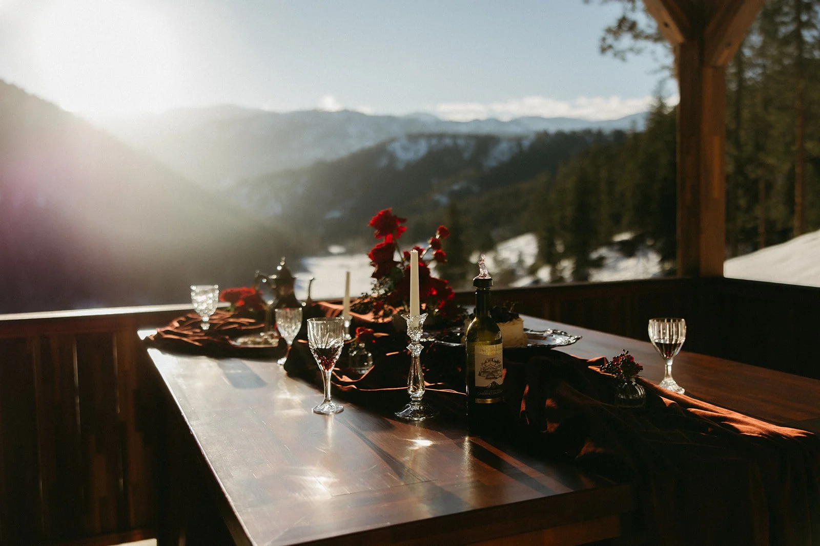 A table set for a meal with wine glasses, a bottle of wine, candlesticks, and a floral centerpiece on a wooden deck with a mountain and forest view in the background.