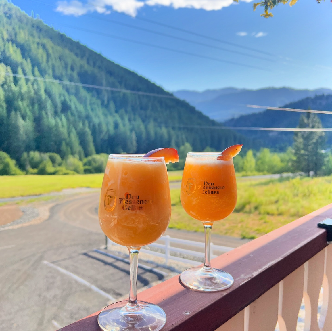 Two glasses of orange-colored drink garnished with orange slices on a balcony railing with a backdrop of green mountains and a blue sky.
