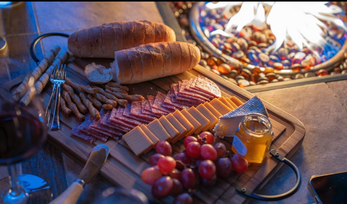 Charcuterie board with bread, sliced meats, cheeses, grapes, honey jar, and a cheese knife, set on a table near a fire pit.