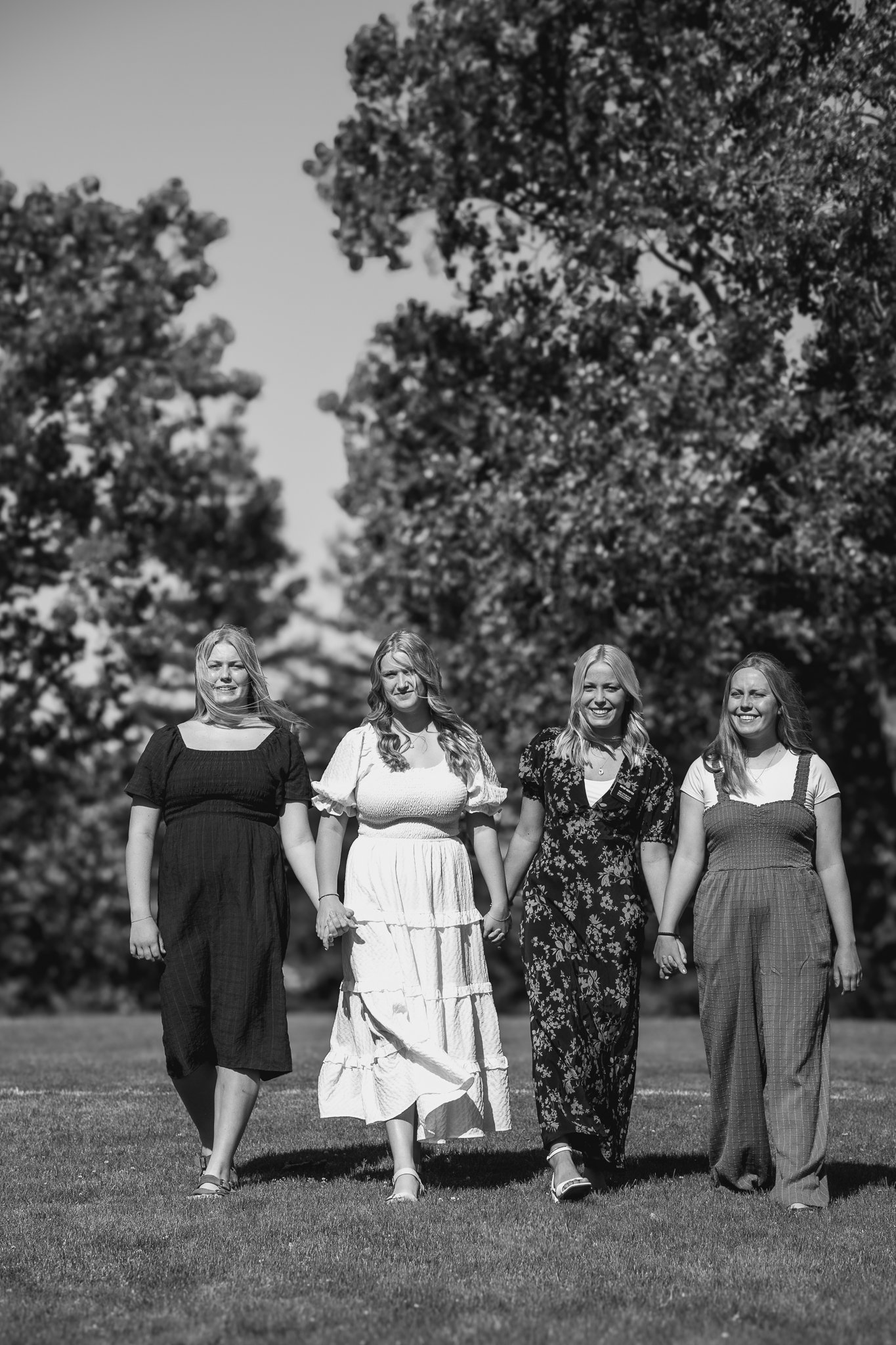 Four women walking hand in hand on grass in a park, wearing dresses and smiling, with trees in the background, black and white photograph.