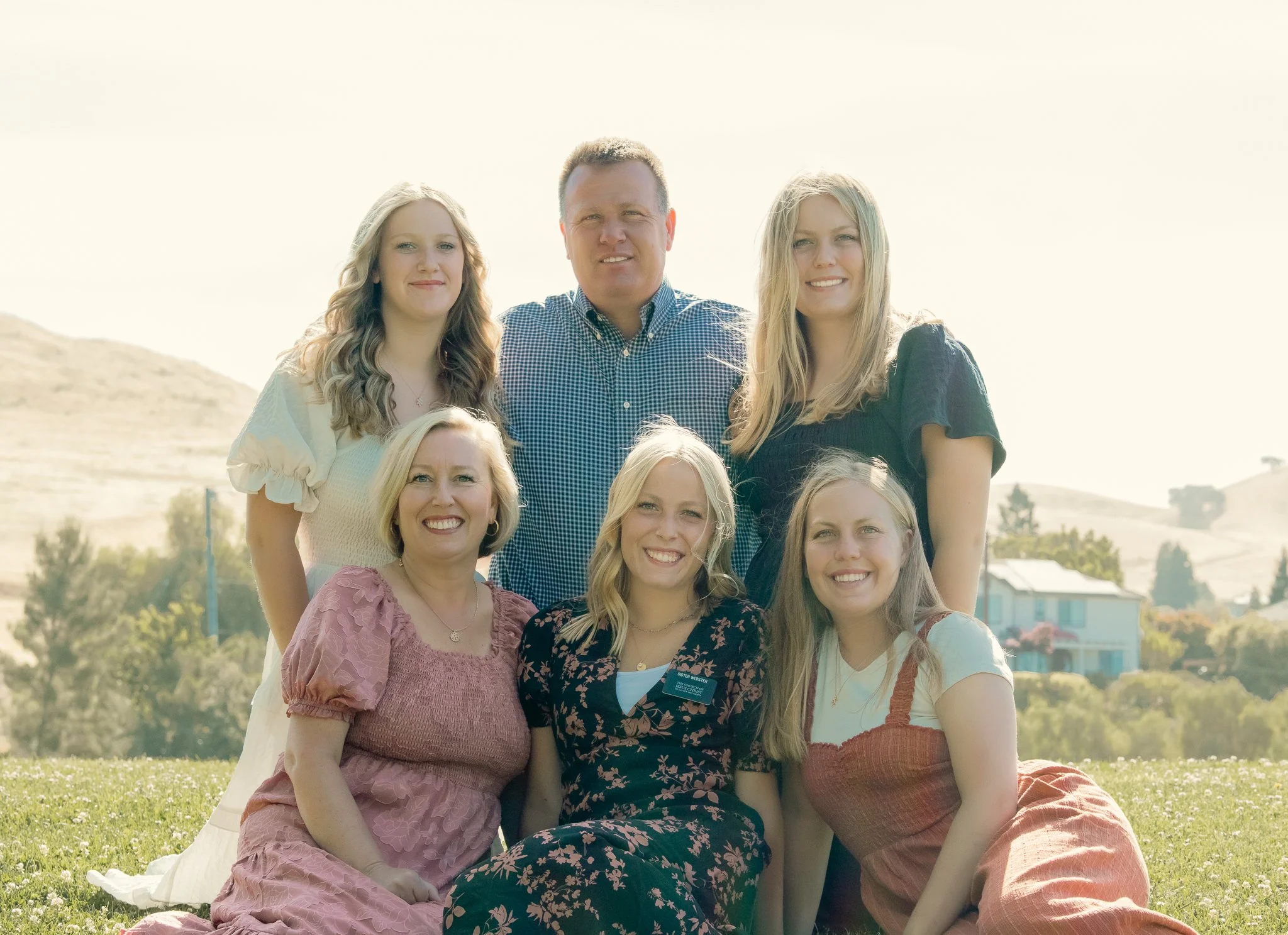 Group of six people, outdoors, in casual clothing, smiling, with rolling hills and houses in the background.
