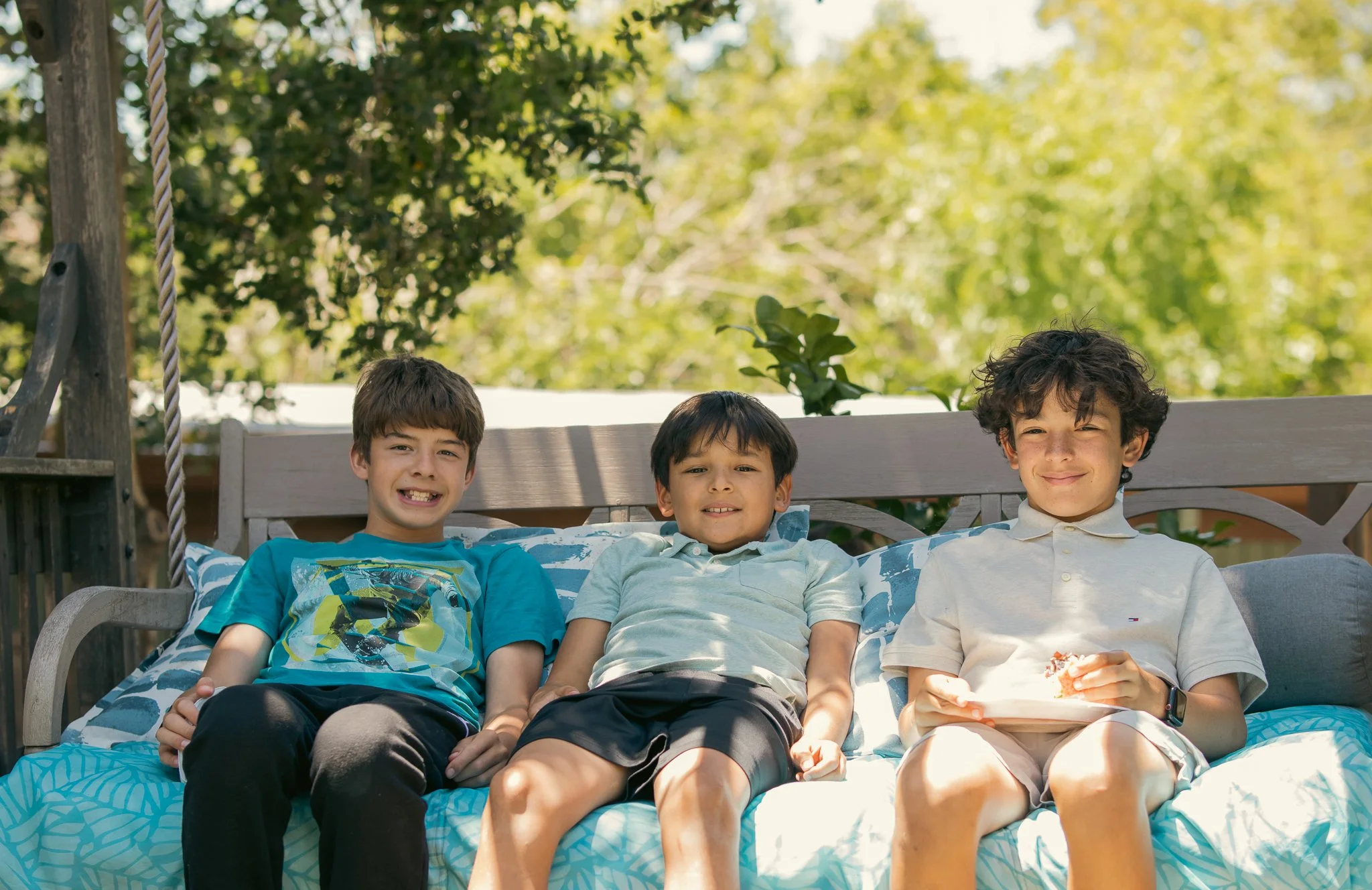 Three boys sitting on an outdoor swing with blue cushions, smiling at the camera with trees in the background.