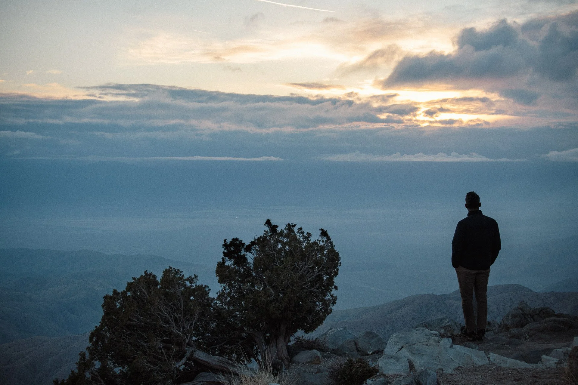 Silhouette of a person standing on a mountain overlook, watching a cloudy sunset; trees and rocky terrain in the foreground.