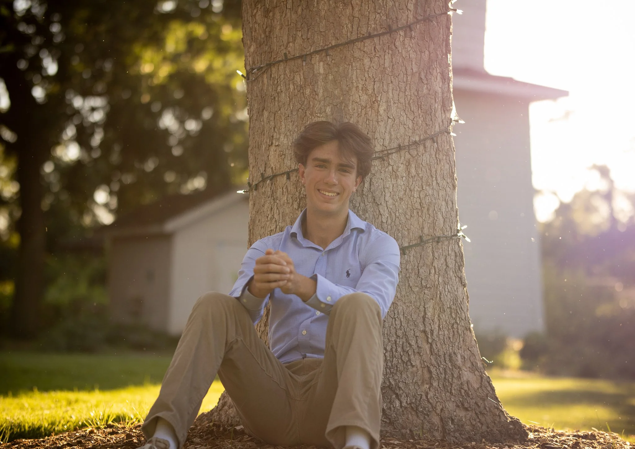 Young man in blue shirt and khaki pants sitting by a tree in a sunny outdoor setting.