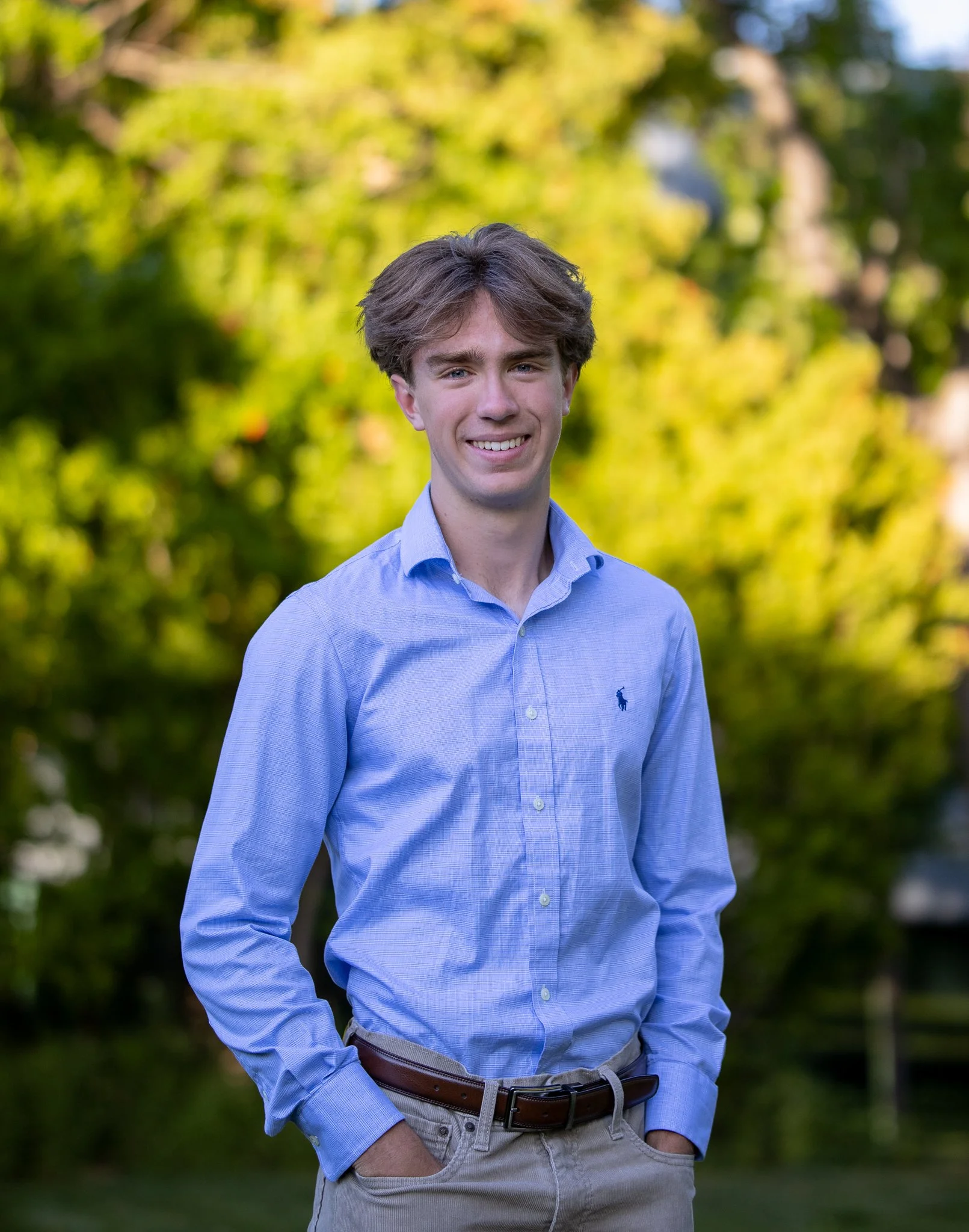 Young man in blue shirt standing outdoors with green foliage background.