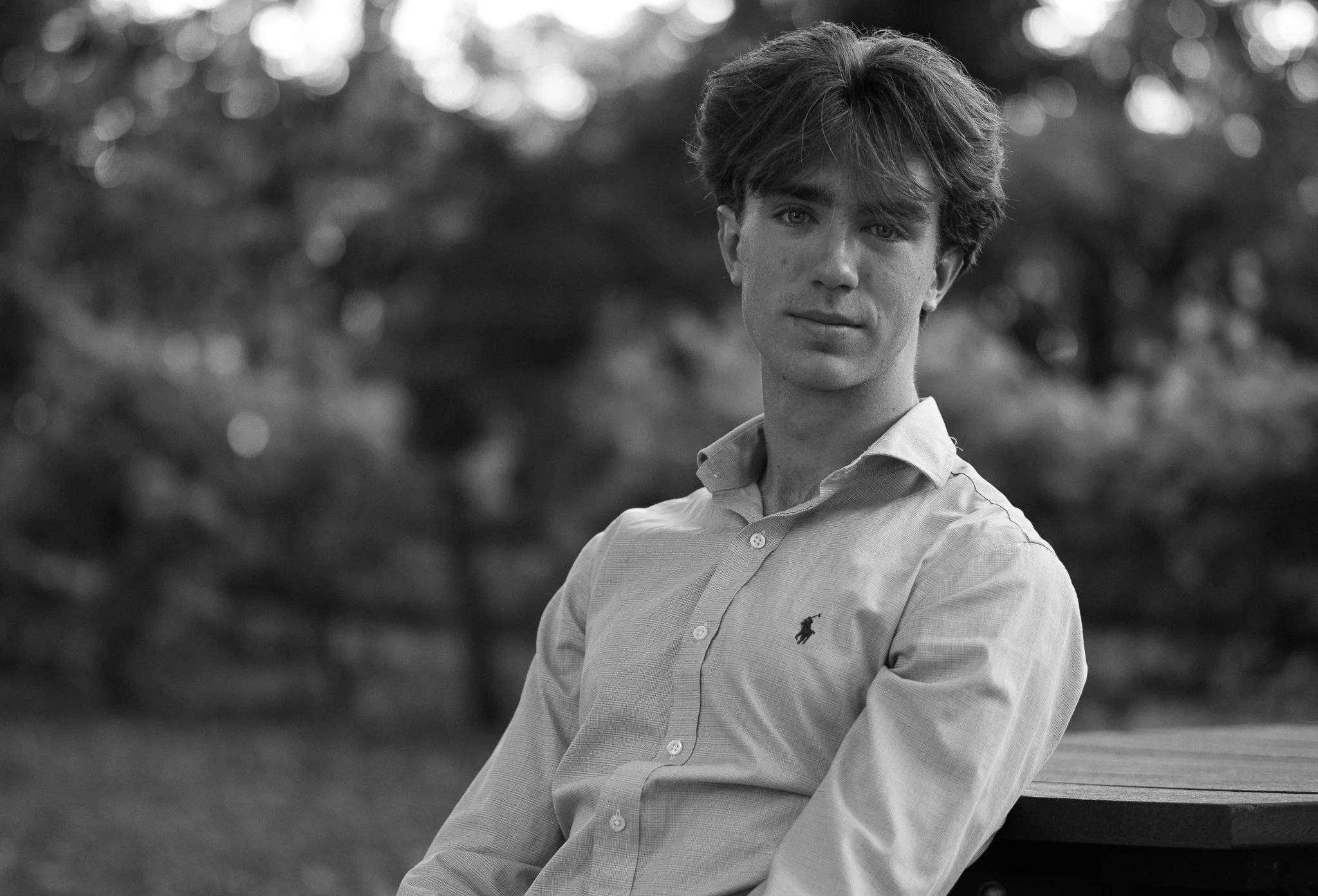 Black and white portrait of a person with medium-length hair in a button-up shirt, sitting outdoors.