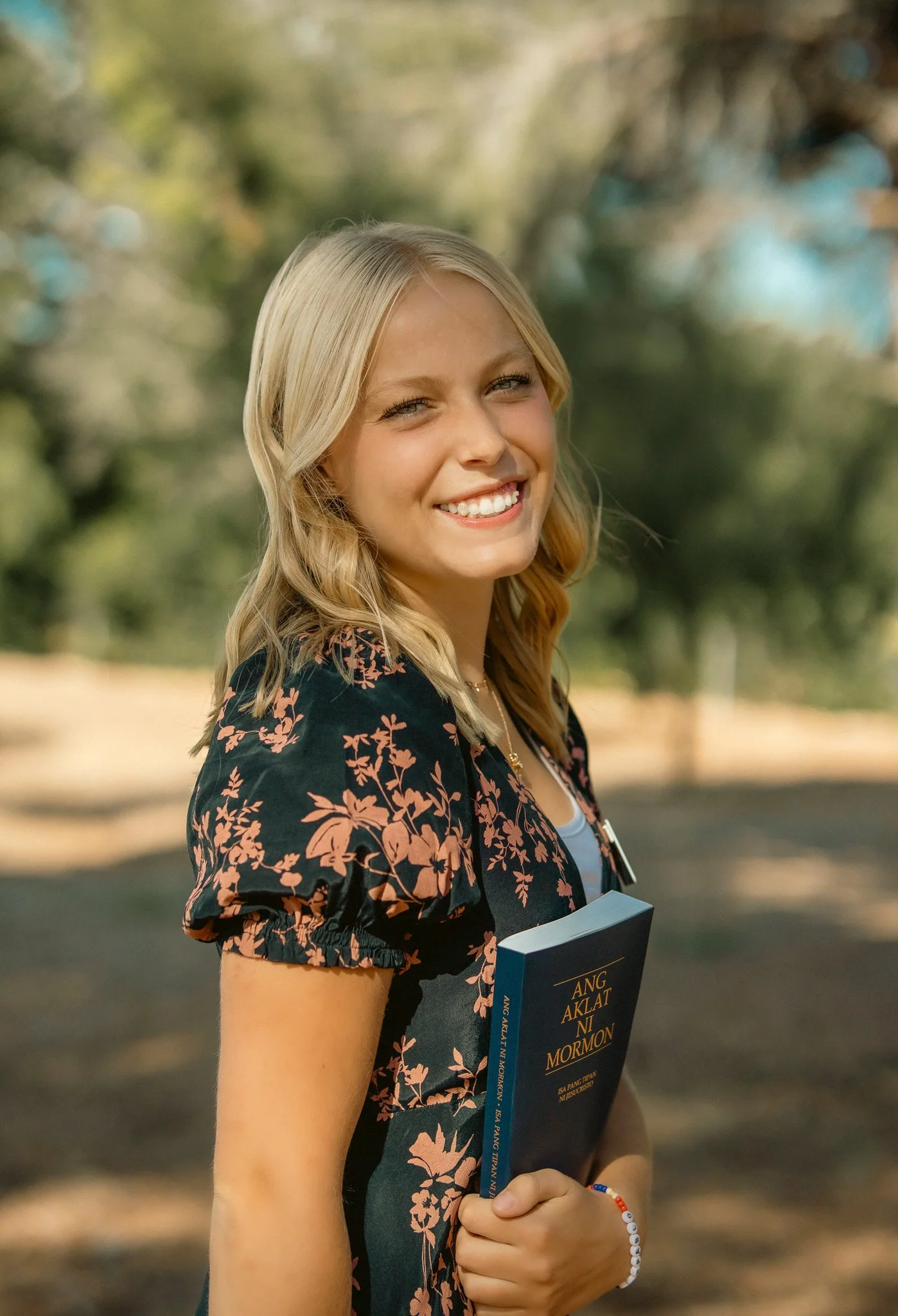 Smiling woman holding "Ang Aklat Ni Mormon" book outdoors.