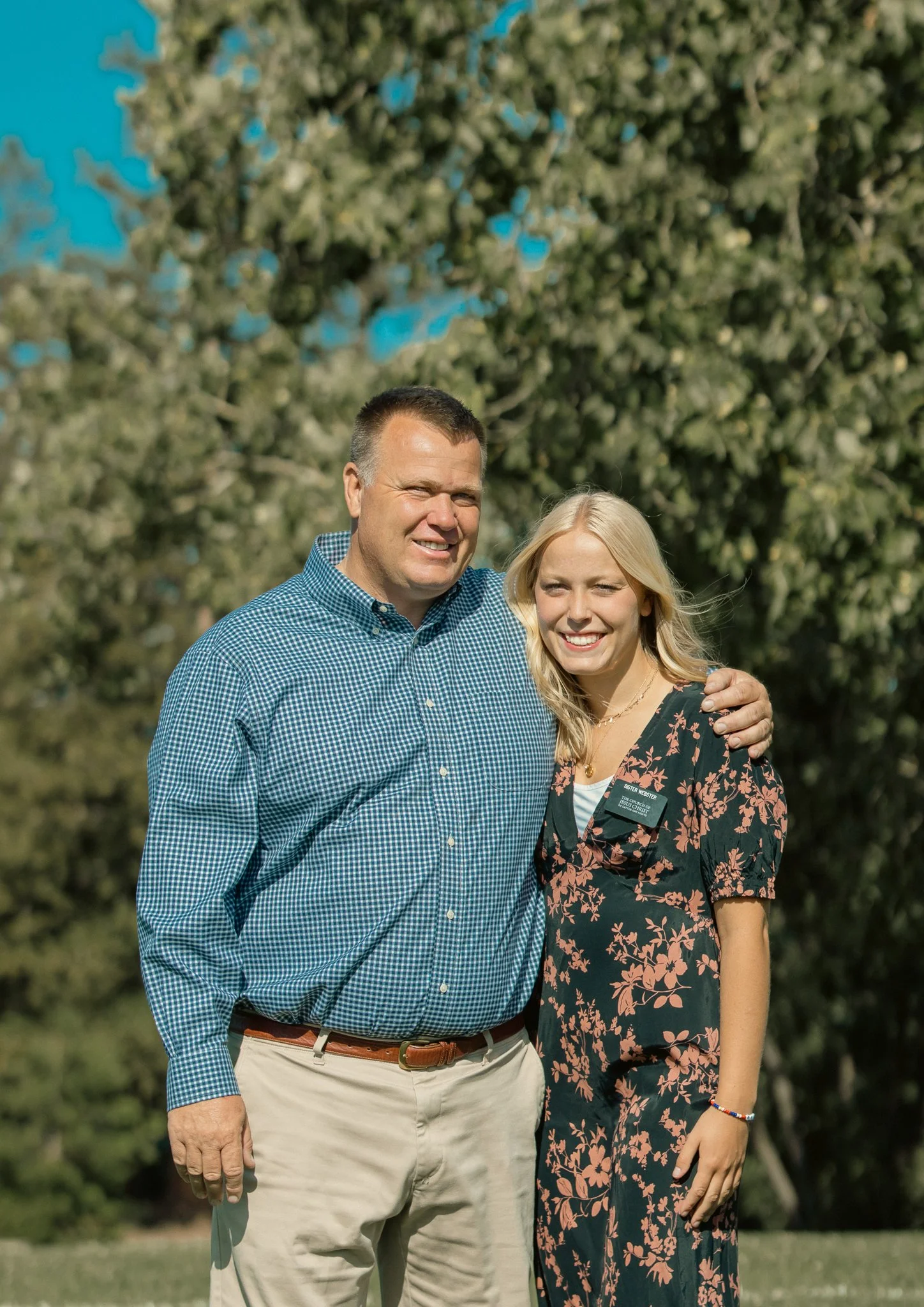 A man and a woman standing together outdoors, smiling, with green trees in the background.