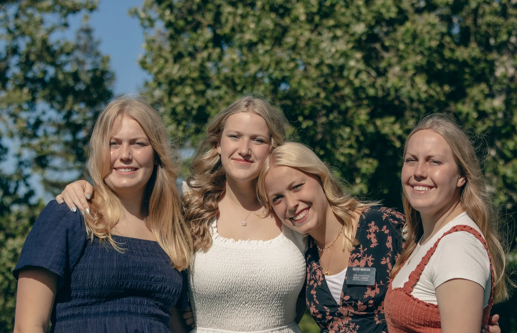 Four young women smiling outdoors, arms around each other, with trees in the background.