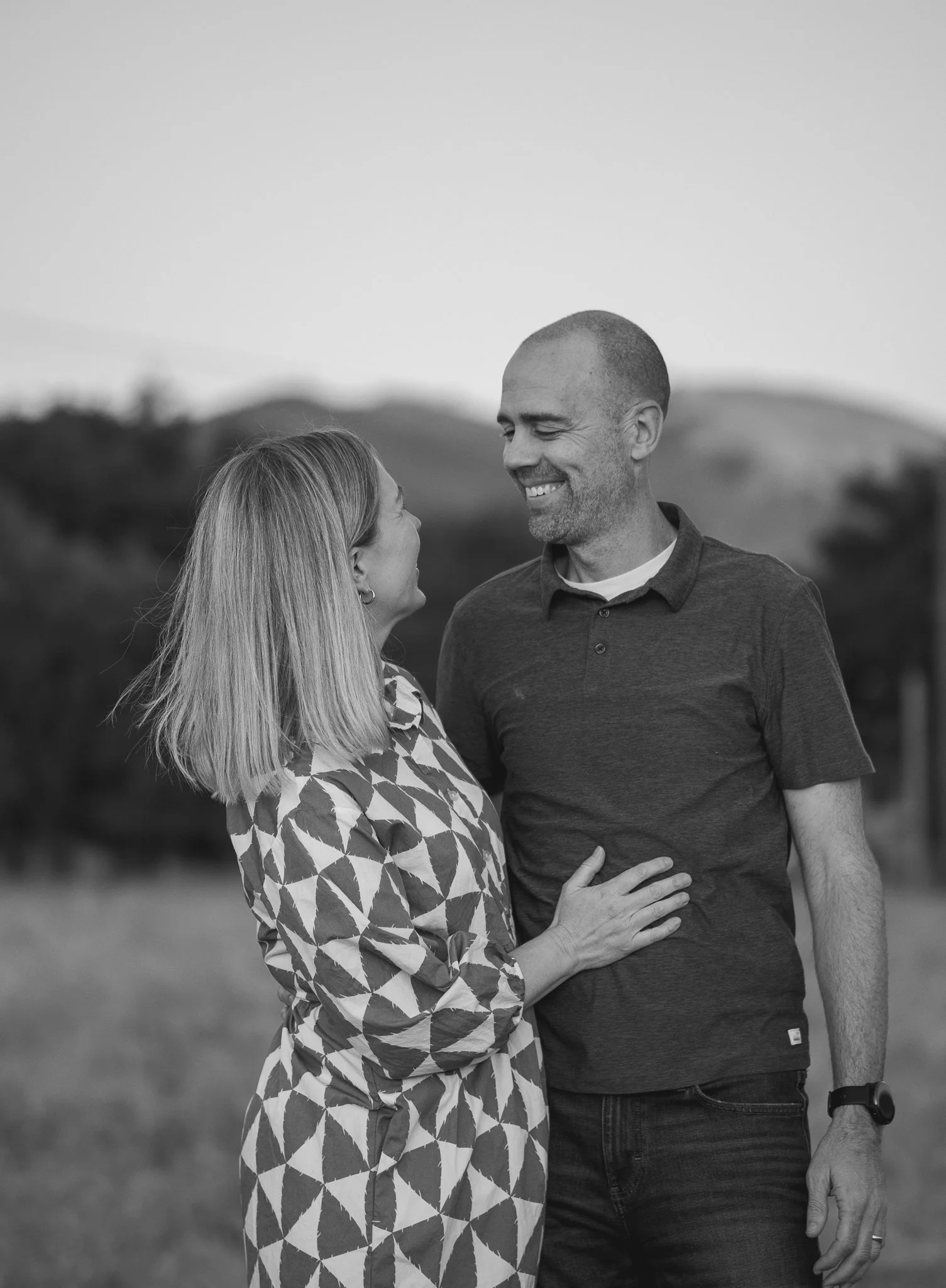 A couple smiling at each other outdoors with blurred trees and hills in the background. The woman is wearing a patterned dress and the man is in a dark shirt.