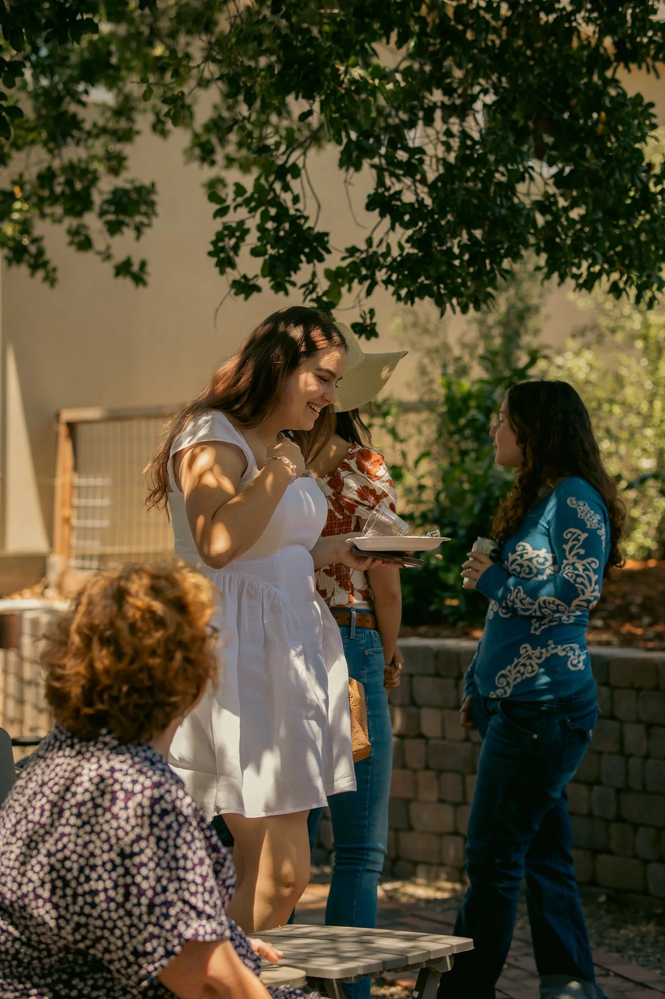 Group of women socializing outdoors, one in a white dress holding a plate, others in casual attire, trees in the background.