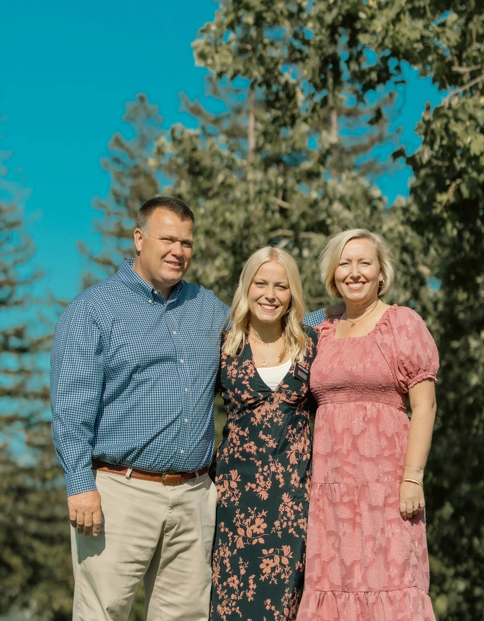 Three people standing outdoors with trees in the background, smiling at the camera. The person on the left is wearing a blue checkered shirt, the person in the middle has long blonde hair and is wearing a floral dress, and the person on the right is wearing a pink dress.