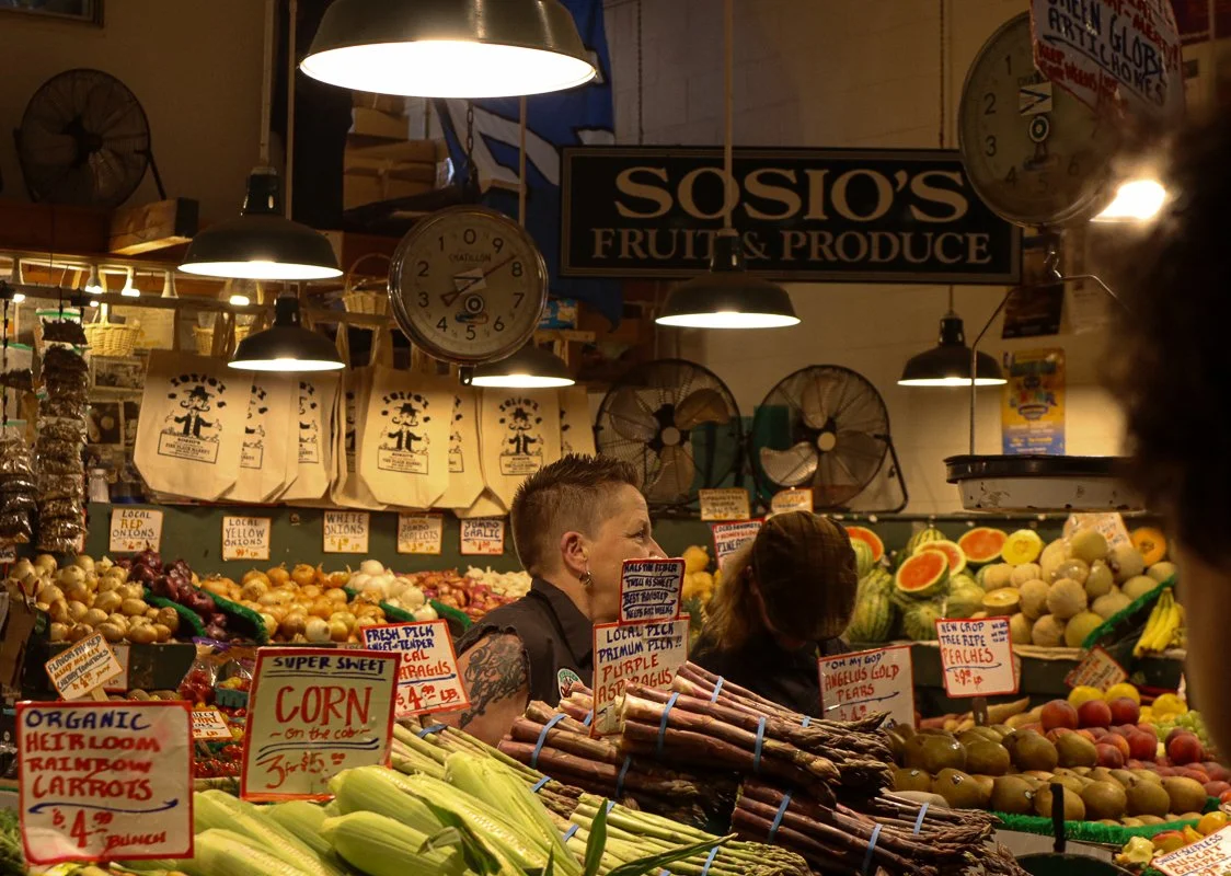 Indoor market stall displaying fresh produce, including corn, asparagus, carrots, and fruits. Signage for Sosio's Fruit & Produce is visible.
