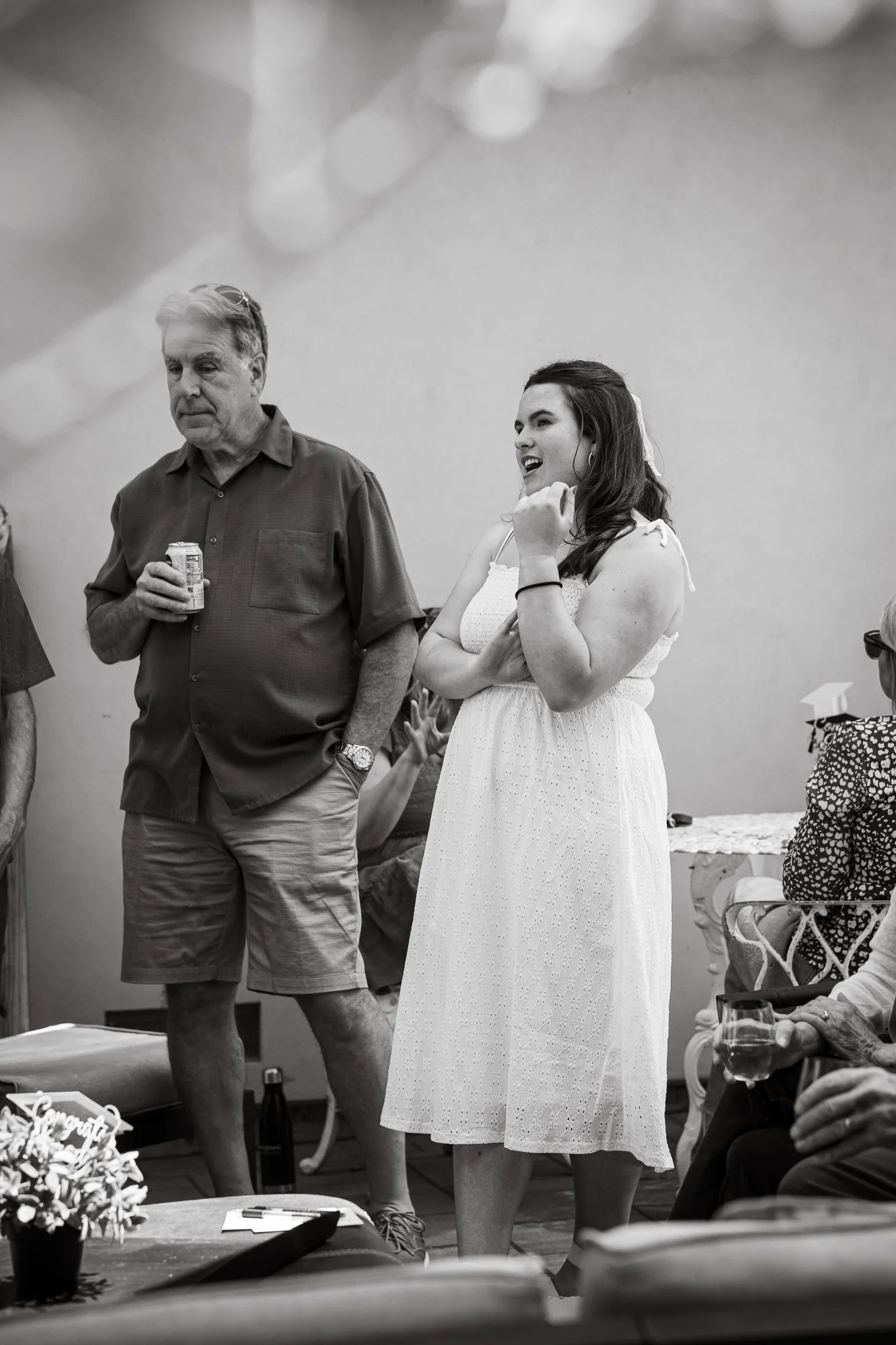 Black and white photo of a group gathering, featuring a man holding a drink and a woman in a white dress talking and smiling.