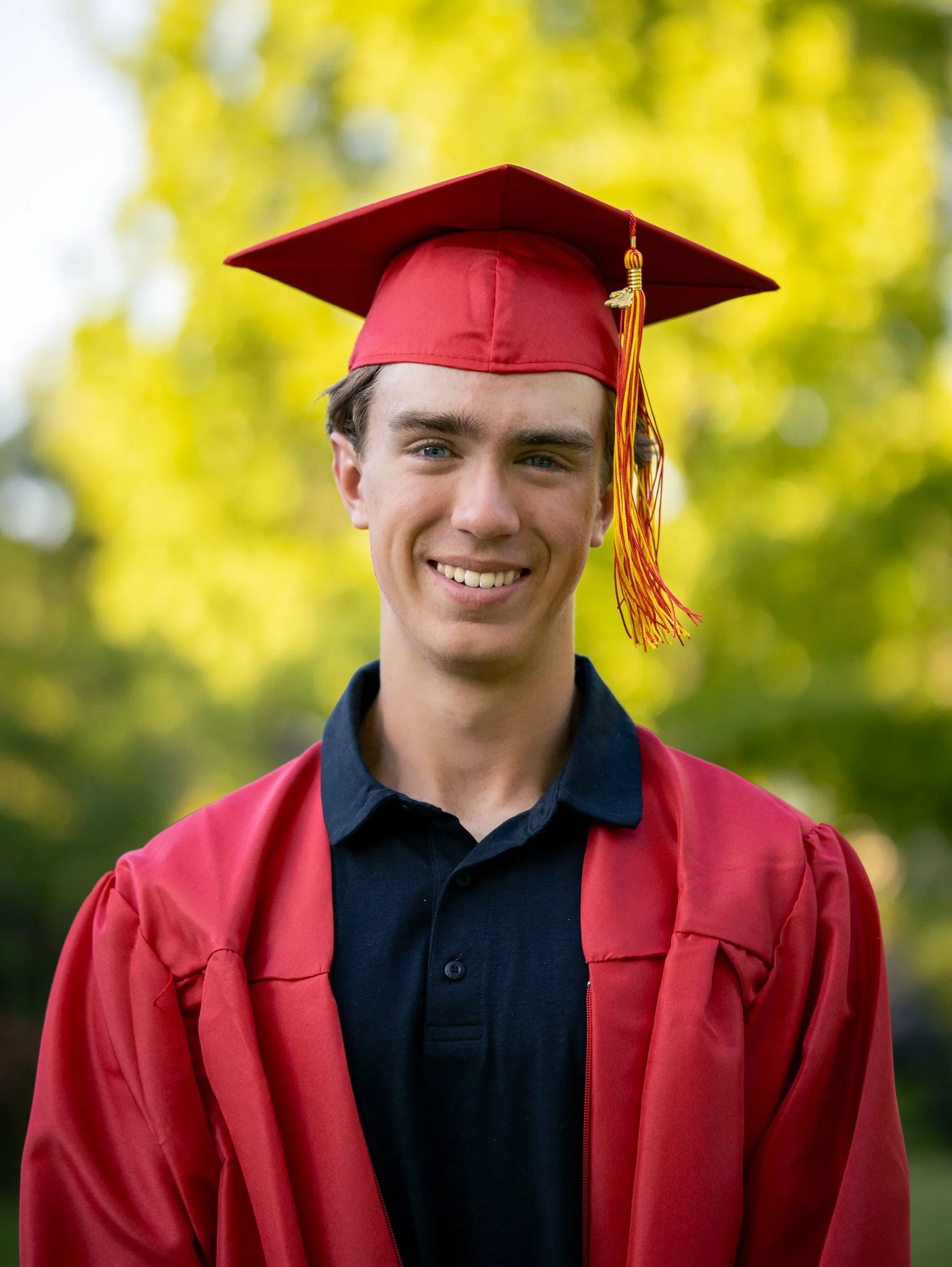 Young man wearing a red graduation cap and gown, smiling outdoors with a blurred green background.