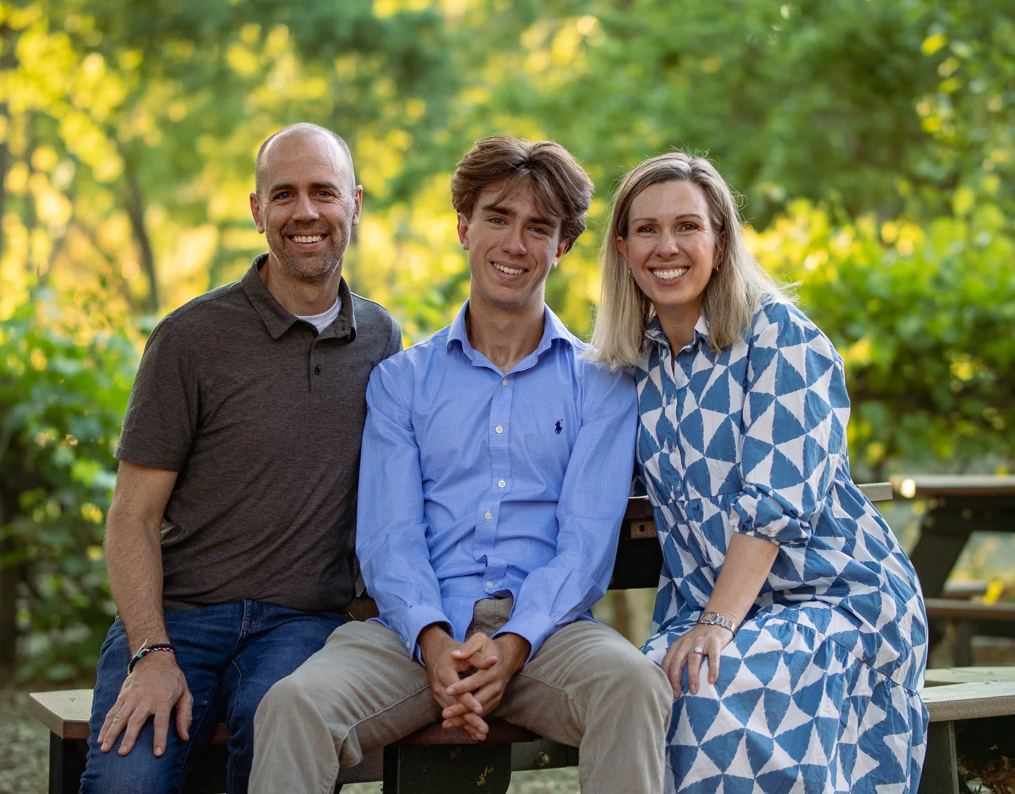 Three people sitting on a bench in a park, smiling, with greenery in the background.