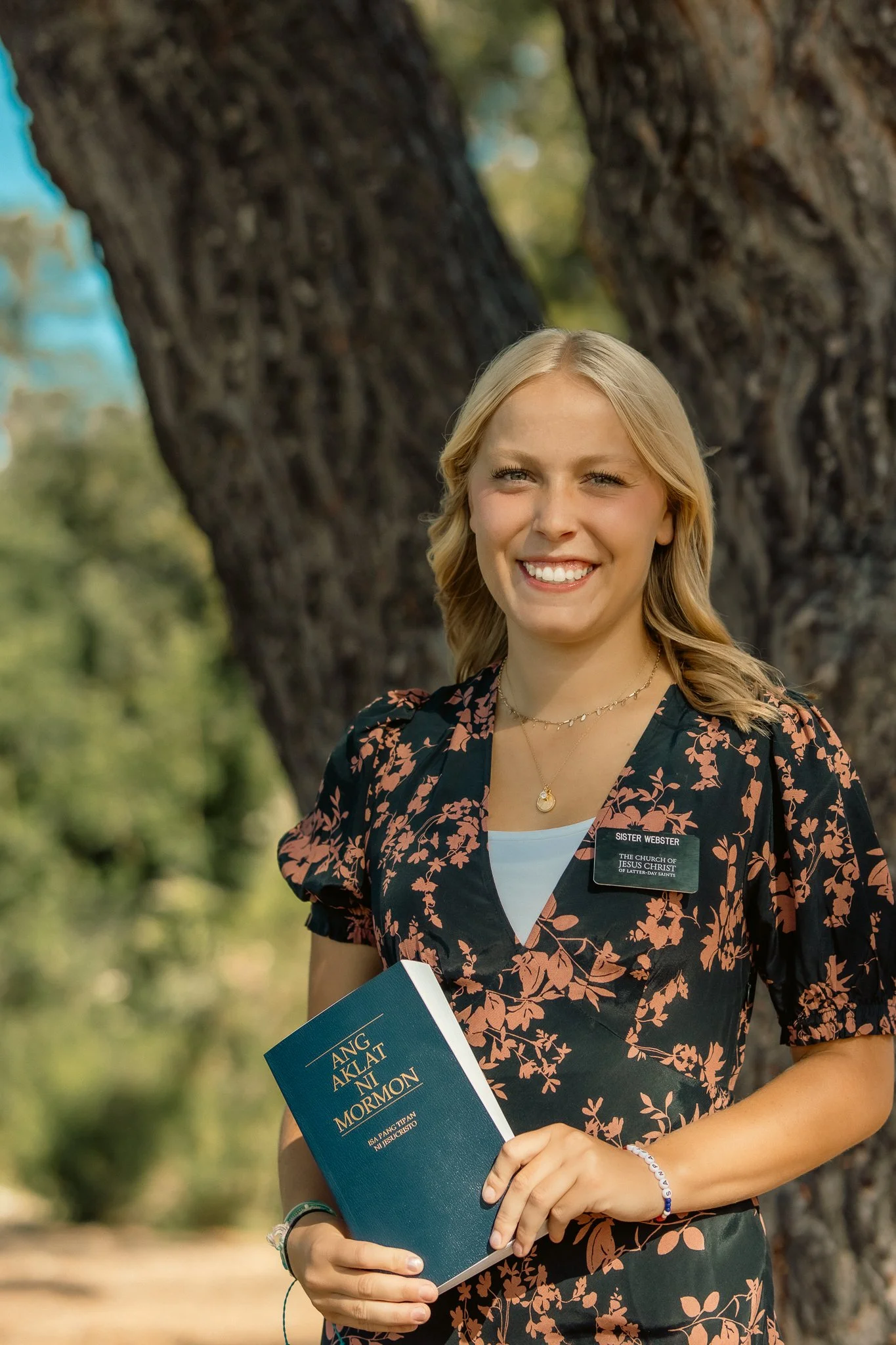 A woman smiling, holding "Ang Aklat ni Mormon" book, wearing a floral dress and a name badge, standing outdoors with trees in the background.