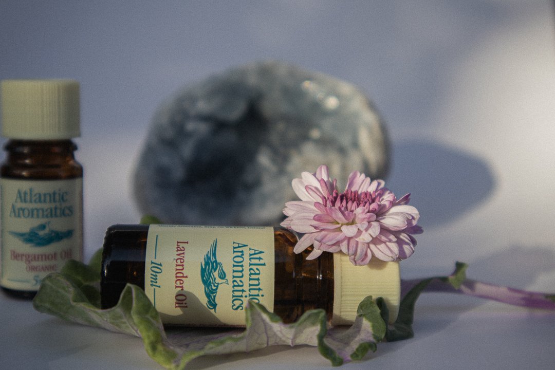 Essential oil bottles labeled "Atlantic Aromatics" with lavender and bergamot oils, accompanied by a pink flower and leaves, set against a blurred stone background.
