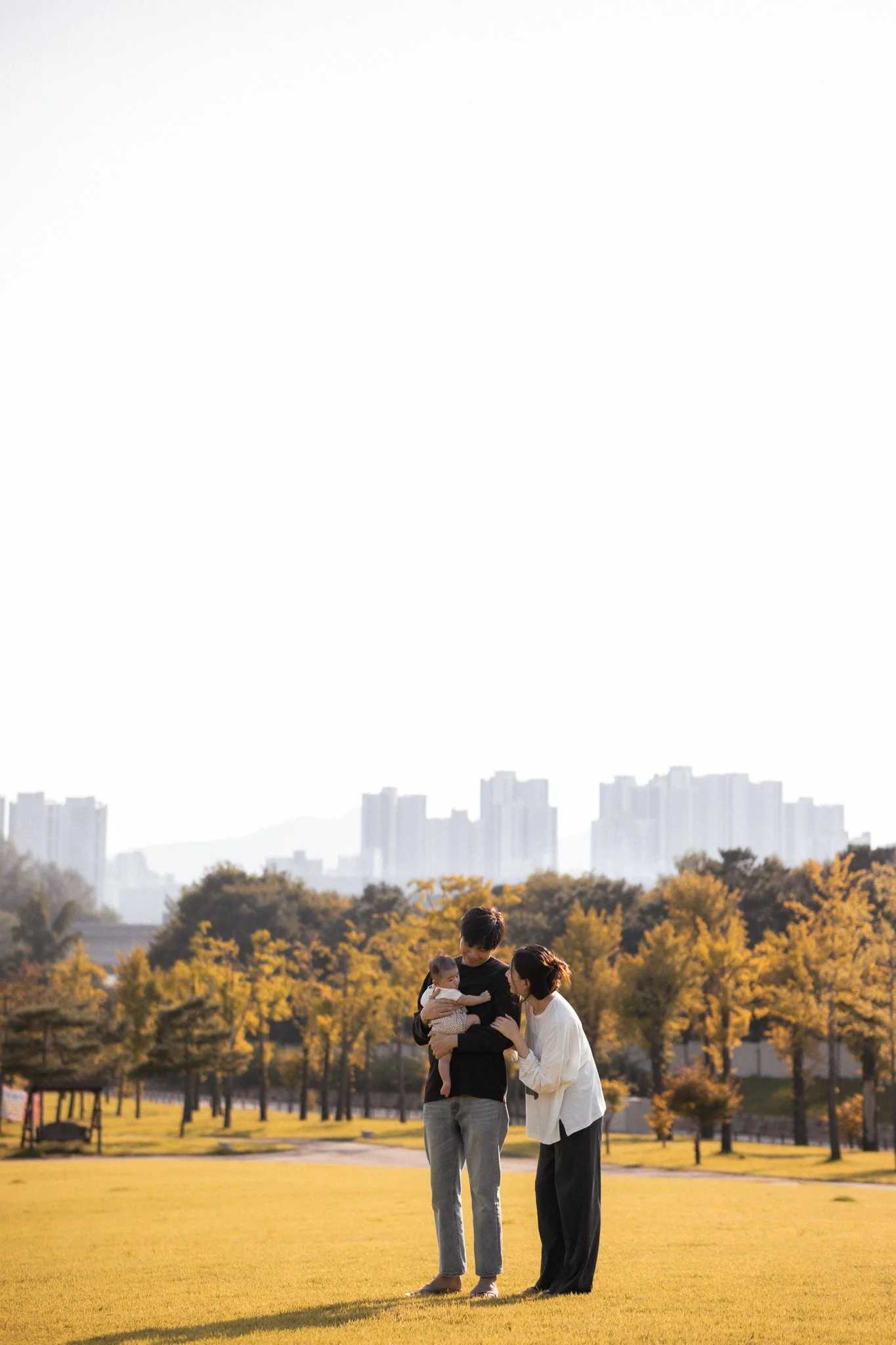 A couple standing on a grassy field holding a baby, with autumn trees and city skyline in the background.