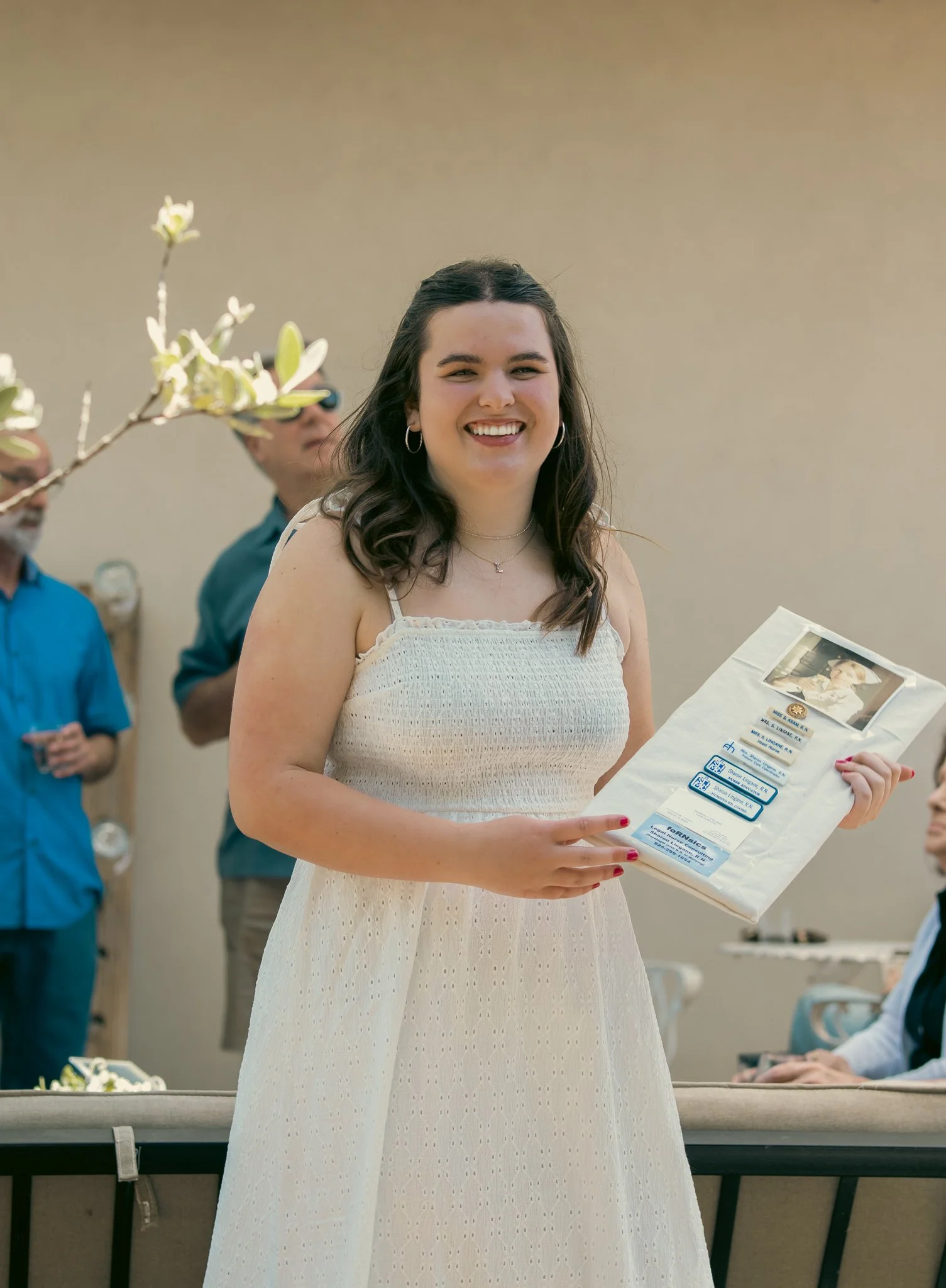 A young woman in a white dress smiling and holding a gift bag at an outdoor gathering, with adults in the background.