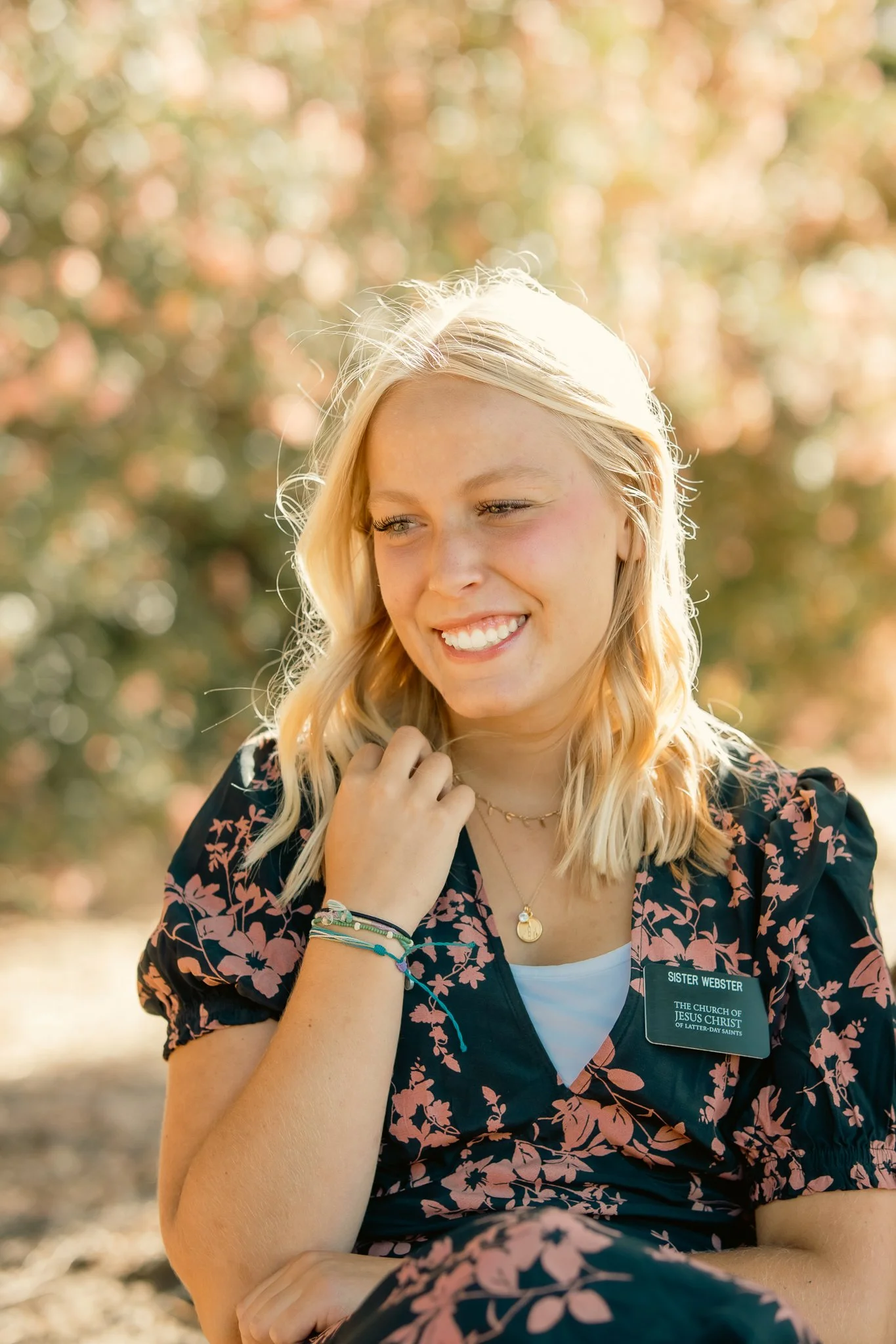 Woman with blonde hair smiling, wearing a floral dress and name tag, sitting outdoors with a blurred background of foliage.