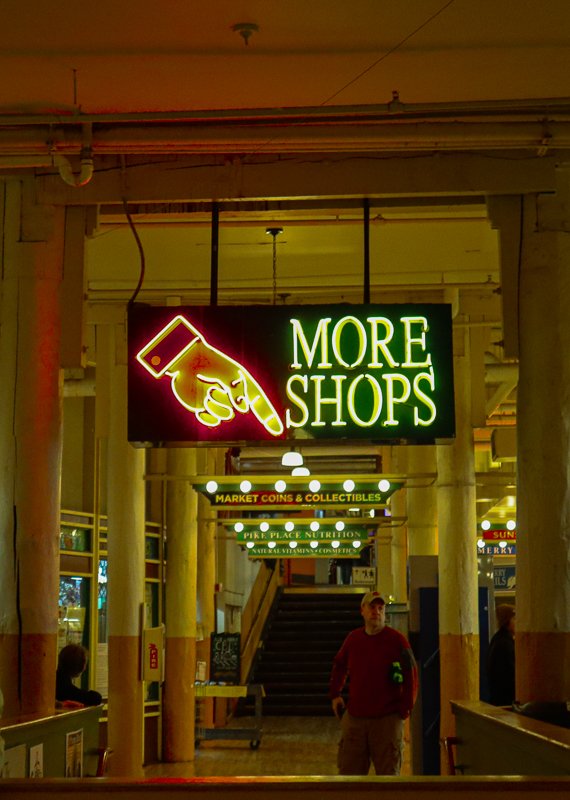Interior of a market with neon sign reading 'More Shops' and an arrow pointing, underscored by various shop signs, including 'Market Coins & Collectibles' and 'Pike Place Nutrition'.
