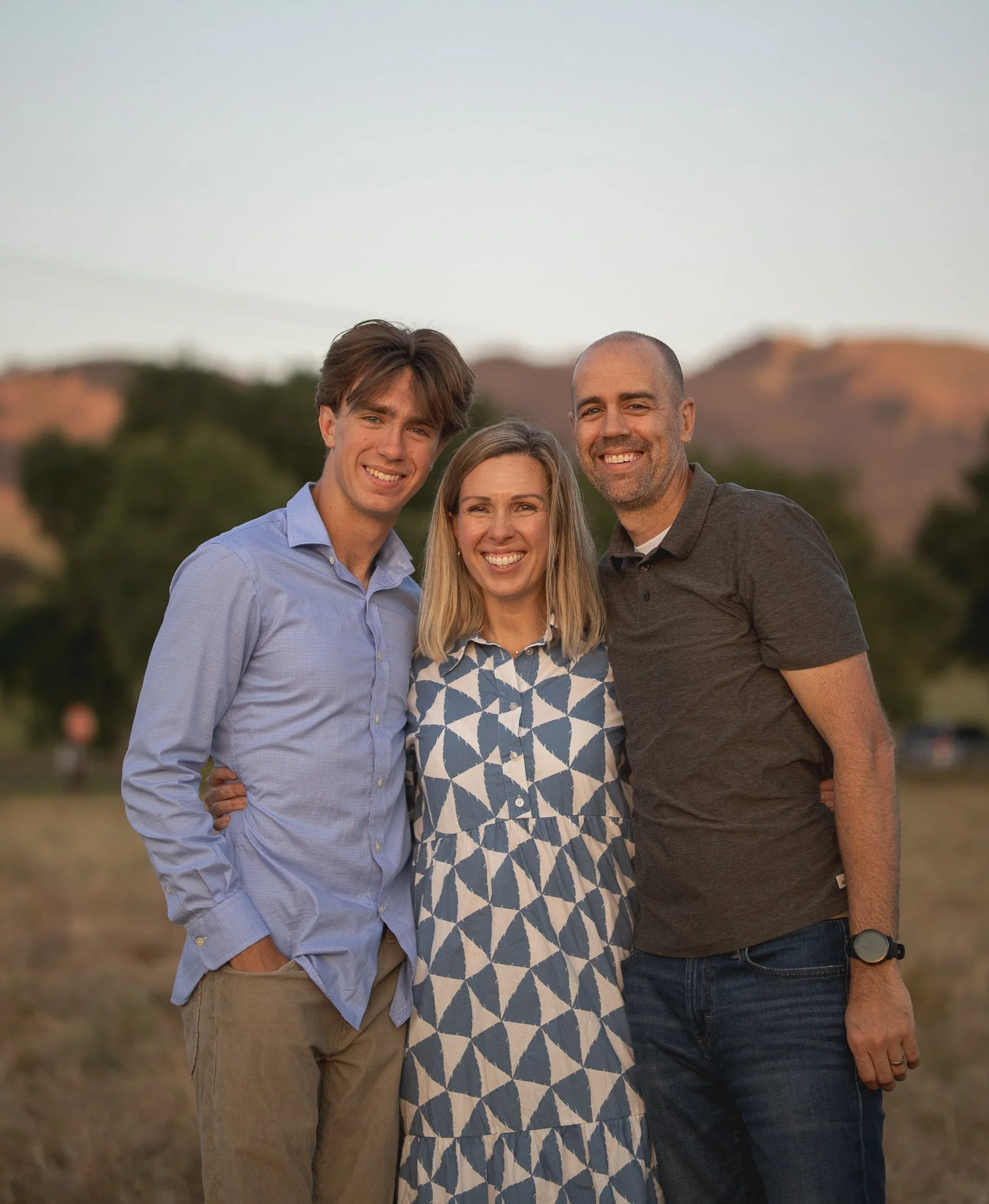 Three people standing outdoors, smiling at the camera. A young man on the left in a blue shirt, a woman in the center in a patterned dress, and a man on the right in a dark polo shirt. Natural background with trees and hills.