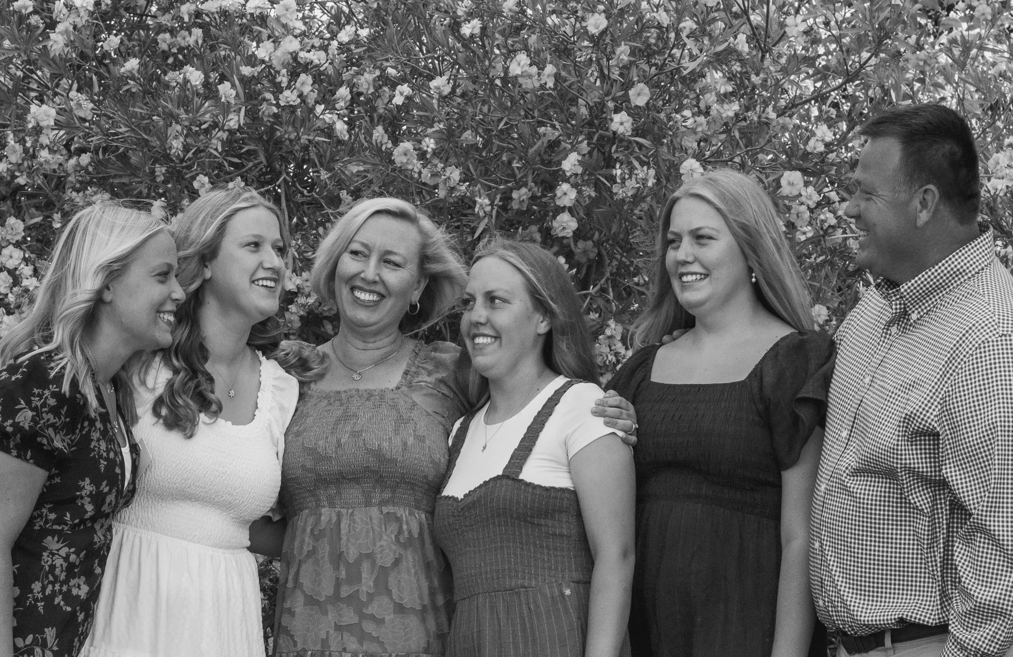 Black and white photo of a group of six people standing together and smiling, with flowering bushes in the background.