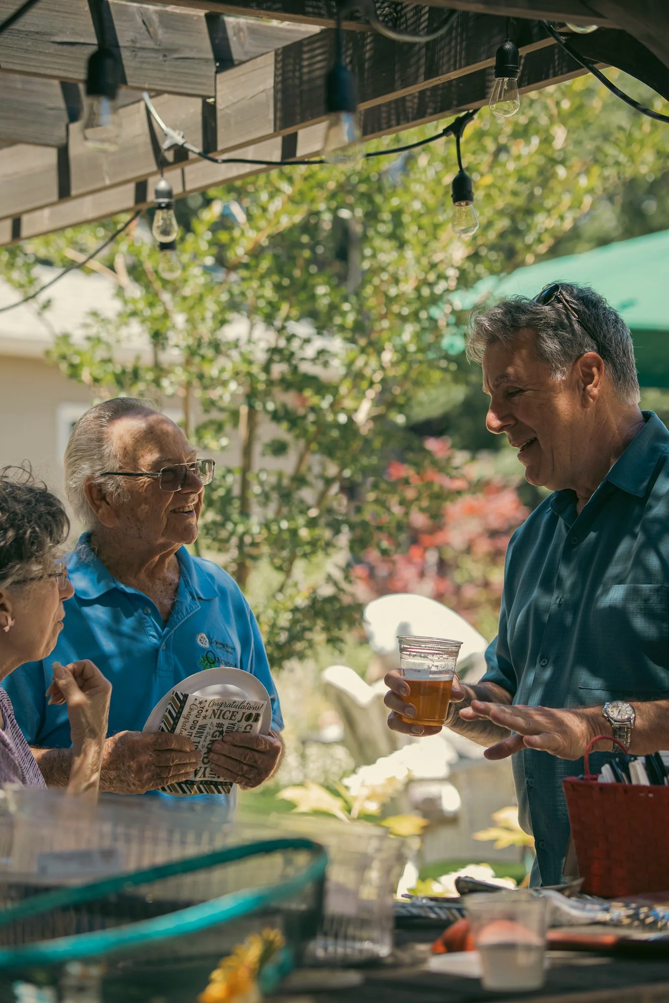 Three people conversing at an outdoor gathering, one holding a drink, under a wooden pergola with string lights, surrounded by greenery.