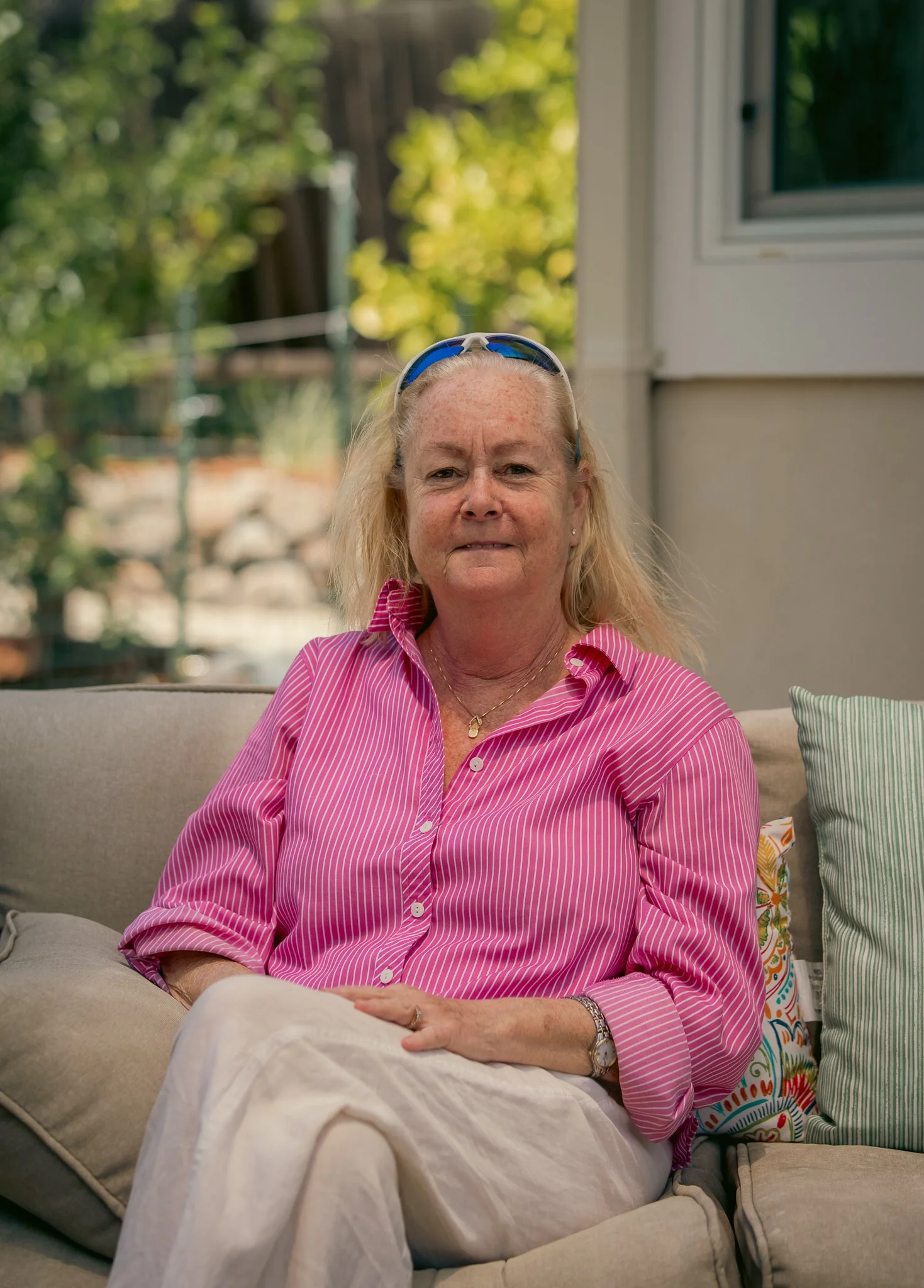A person wearing a pink striped shirt and sunglasses sits on an outdoor sofa with colorful cushions.