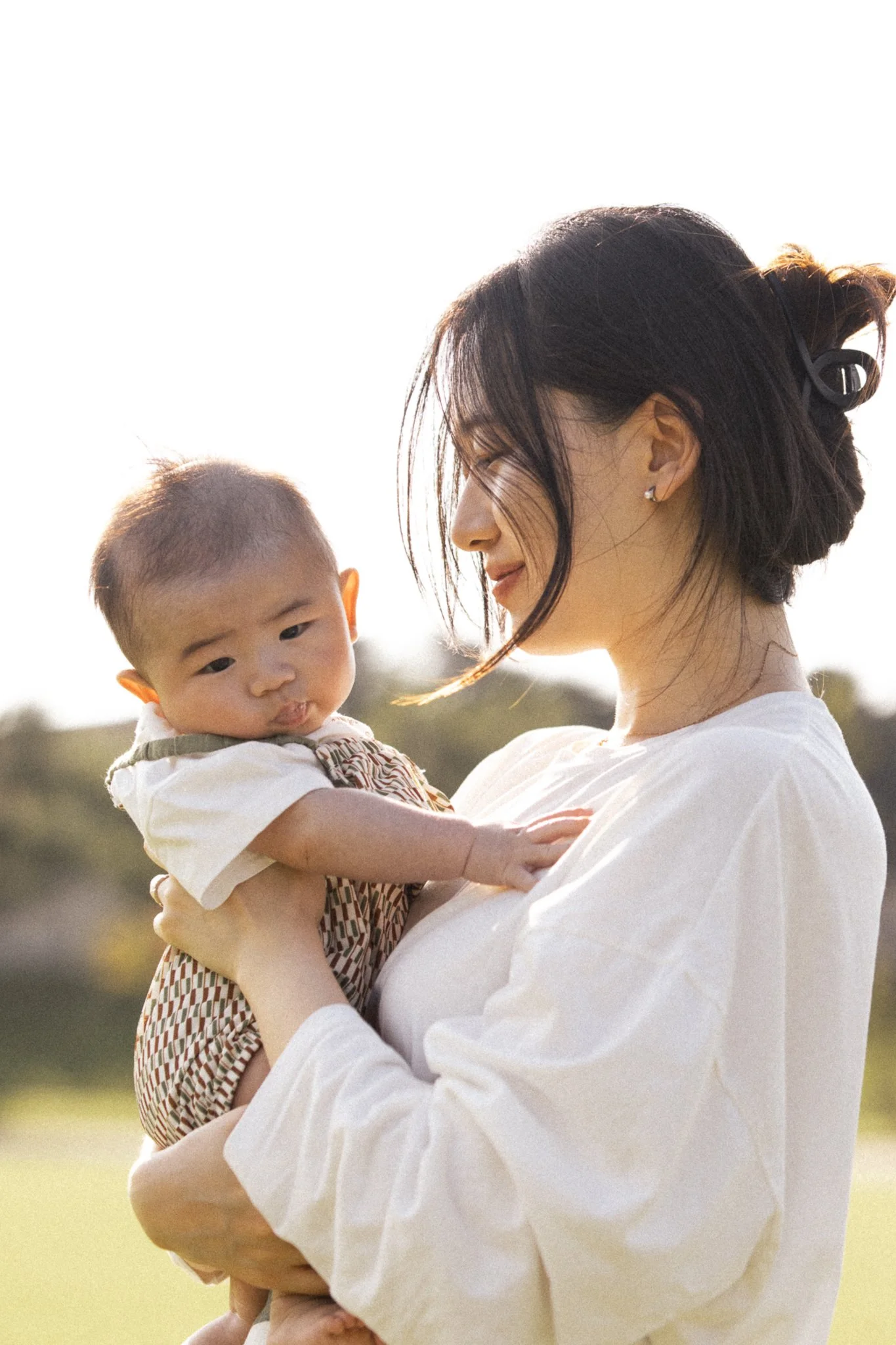 A woman holding a baby outdoors, both looking content and happy under the sunlight.