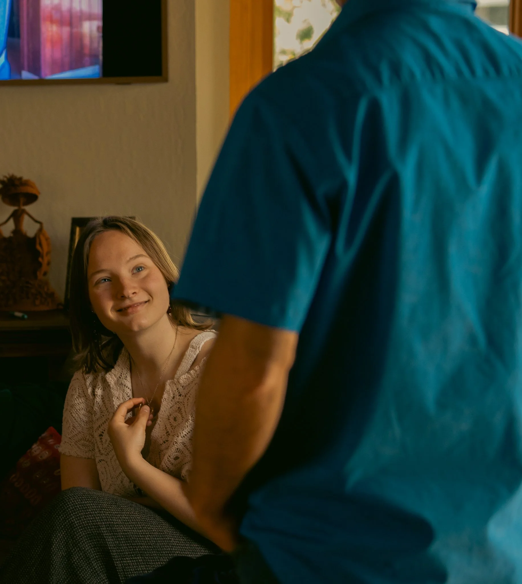 A girl smiling and looking at a person wearing a blue shirt, indoors, with a TV in the background.