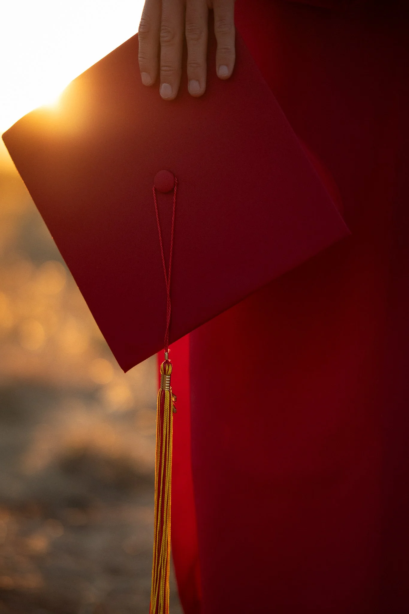 Person holding a red graduation cap with a yellow tassel at sunset.