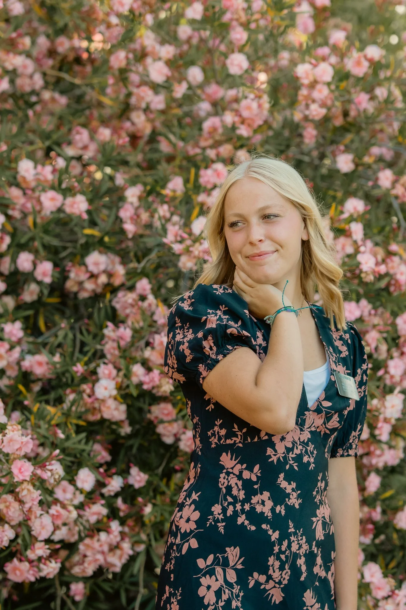 Blonde woman in floral dress standing in front of pink blooming oleander bush, looking thoughtful.
