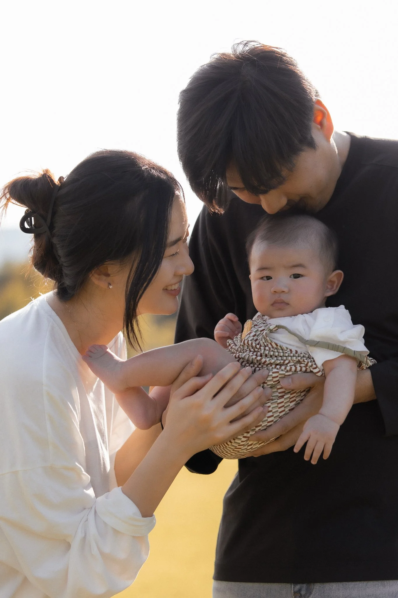 A couple lovingly holding a baby outdoors, with the woman smiling and the man gently kissing the baby's head.
