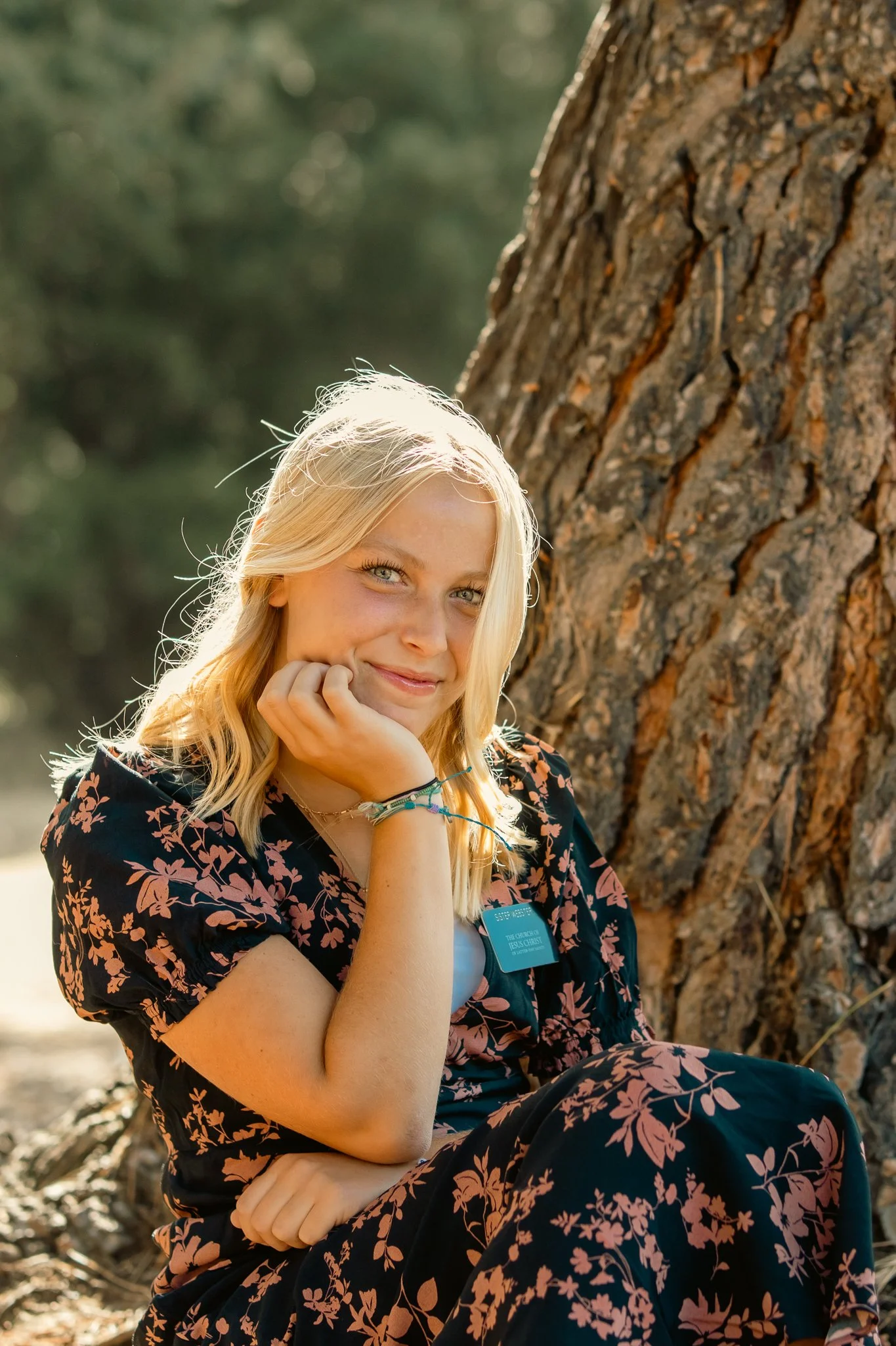 Person with blonde hair wearing a floral dress sitting by a tree, smiling at the camera.
