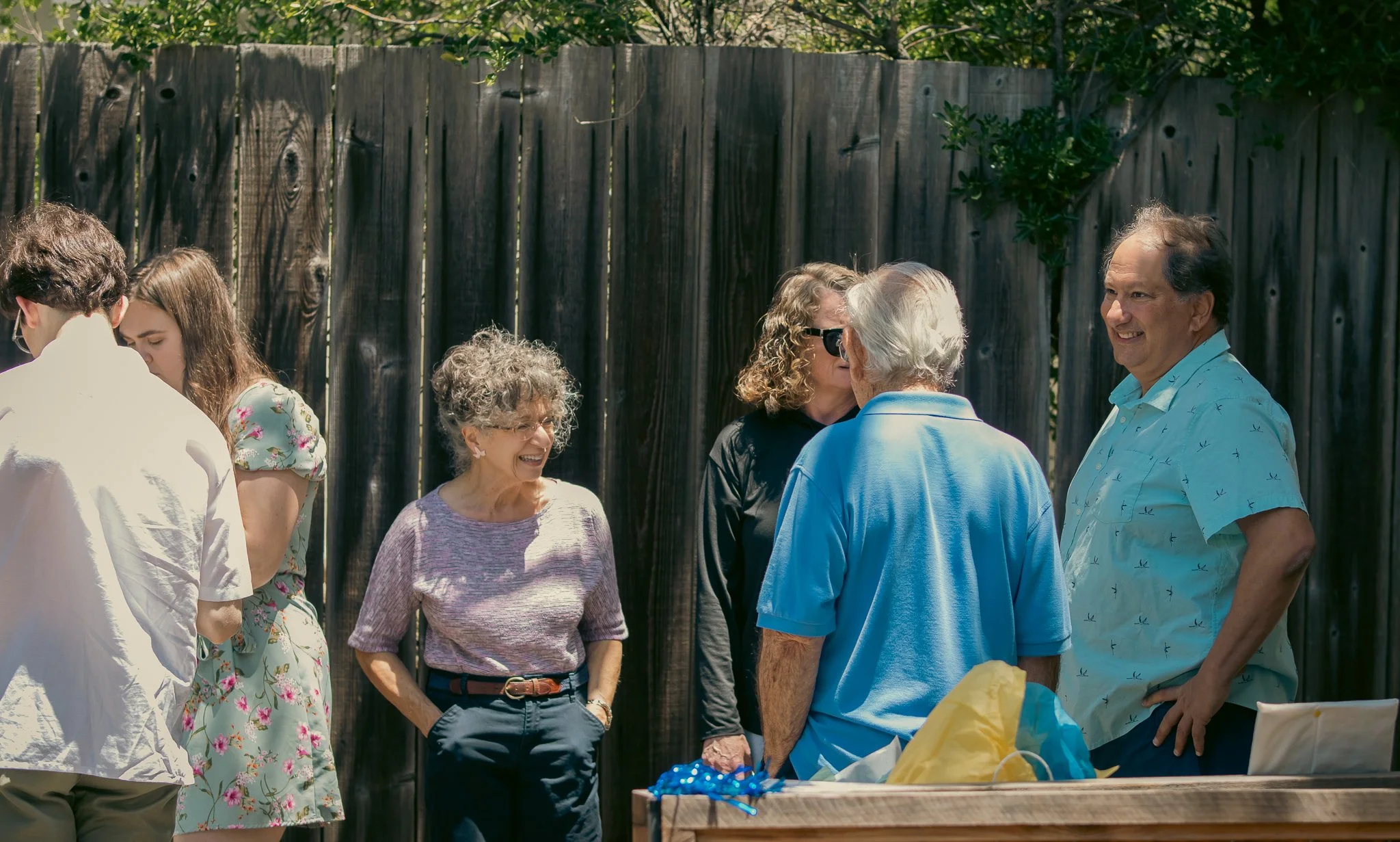 A group of people chatting and smiling at an outdoor gathering near a wooden fence.