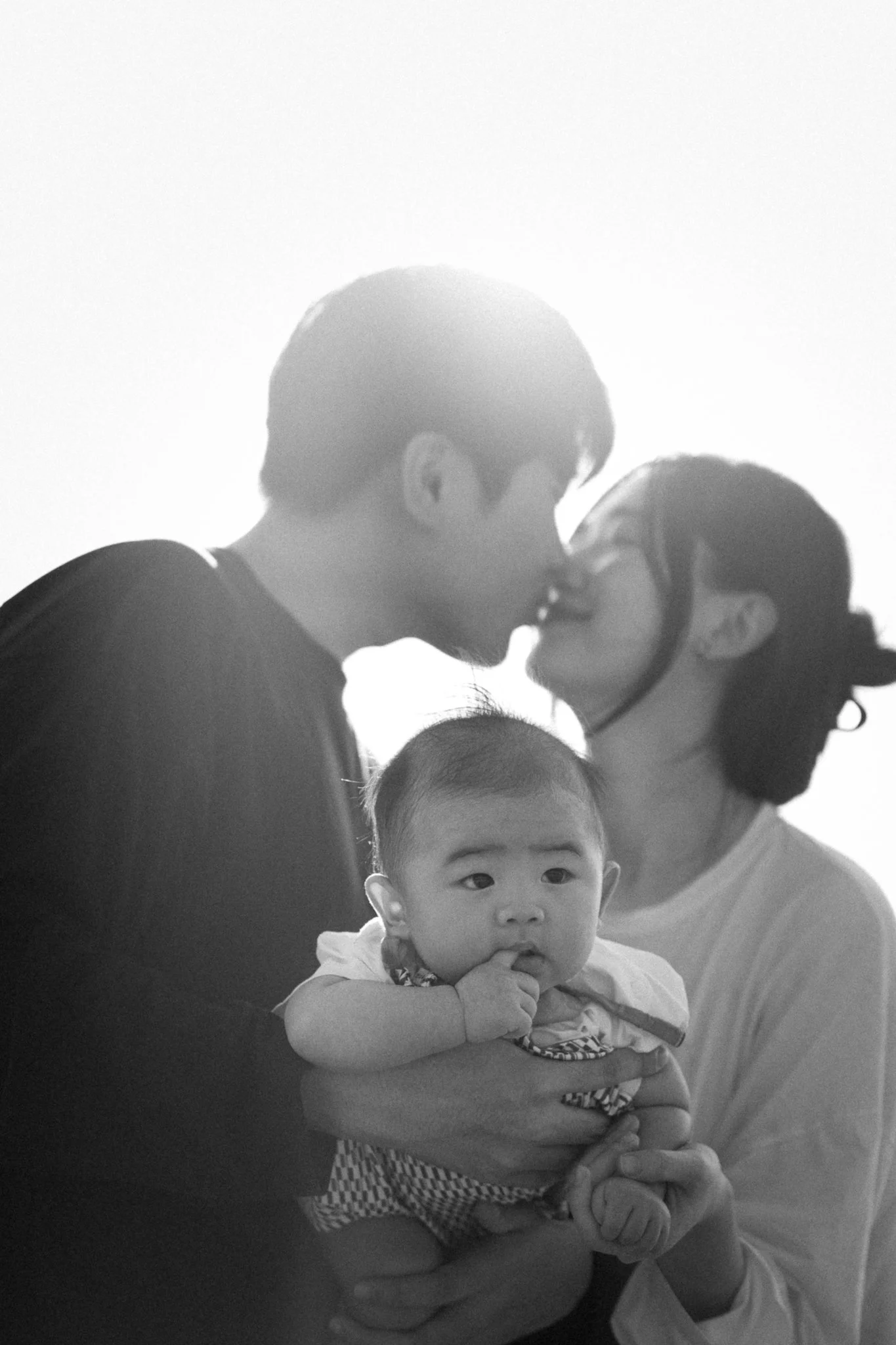Black and white photo of a couple kissing with a baby in the foreground.