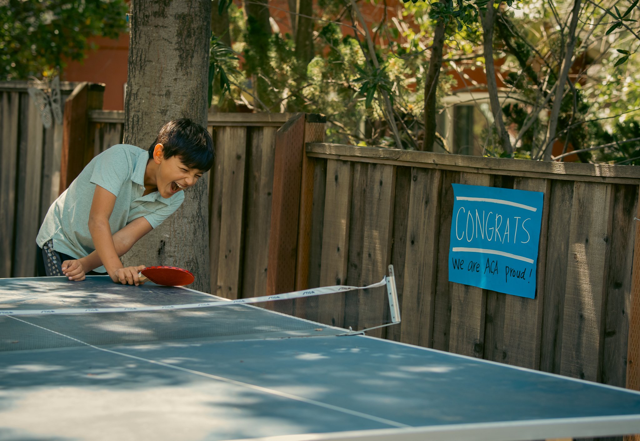 Boy playing table tennis outside, smiling, with a wooden fence and a congratulatory sign in the background.
