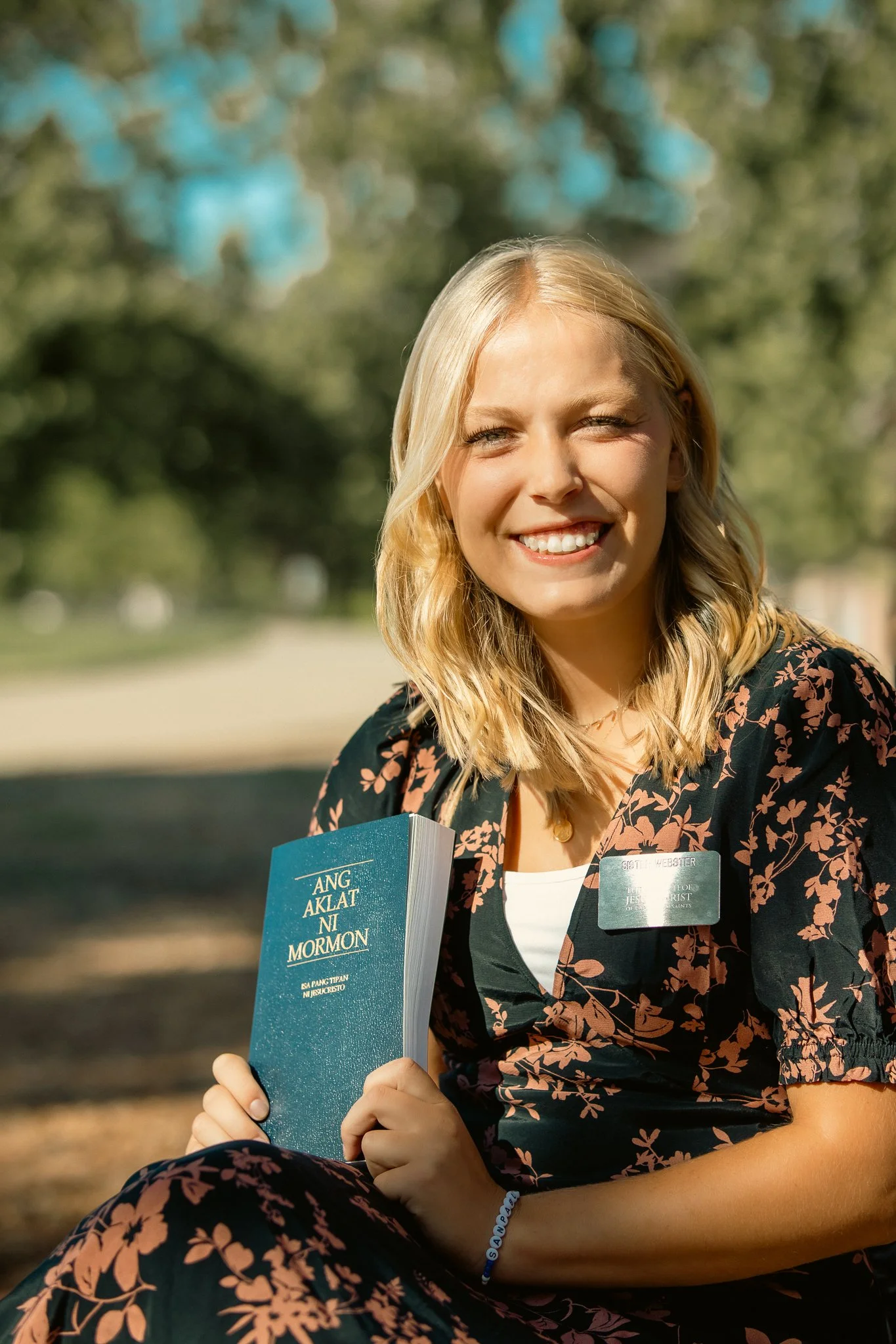 A person sitting outdoors holding "Ang Aklat ni Mormon" book in Filipino, smiling with a name tag on their floral dress, in a park setting.