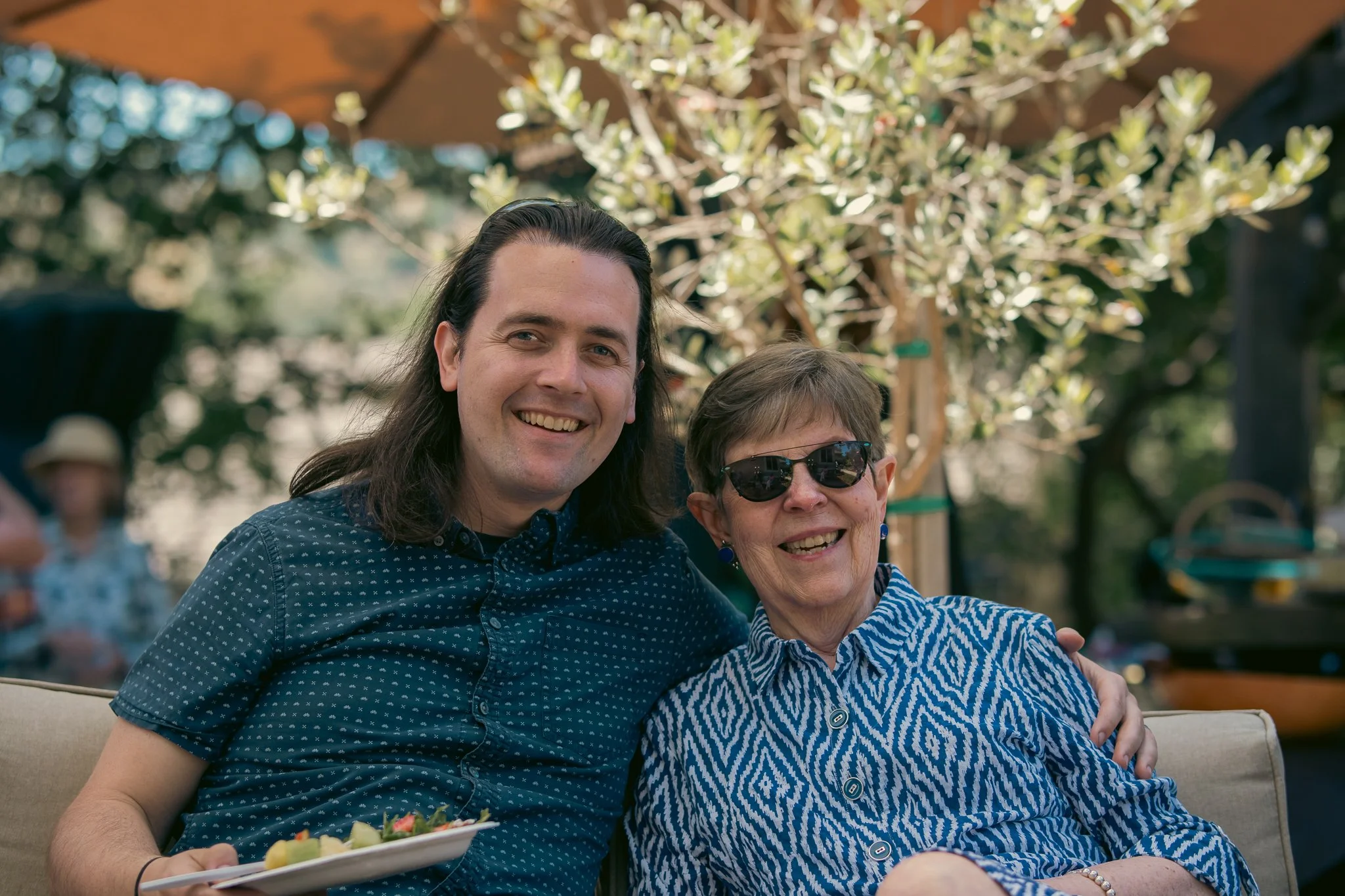 Two people sitting outdoors, smiling. Both are wearing patterned shirts. The person on the left has long hair and the person on the right is wearing sunglasses. There are trees and a canopy in the background.