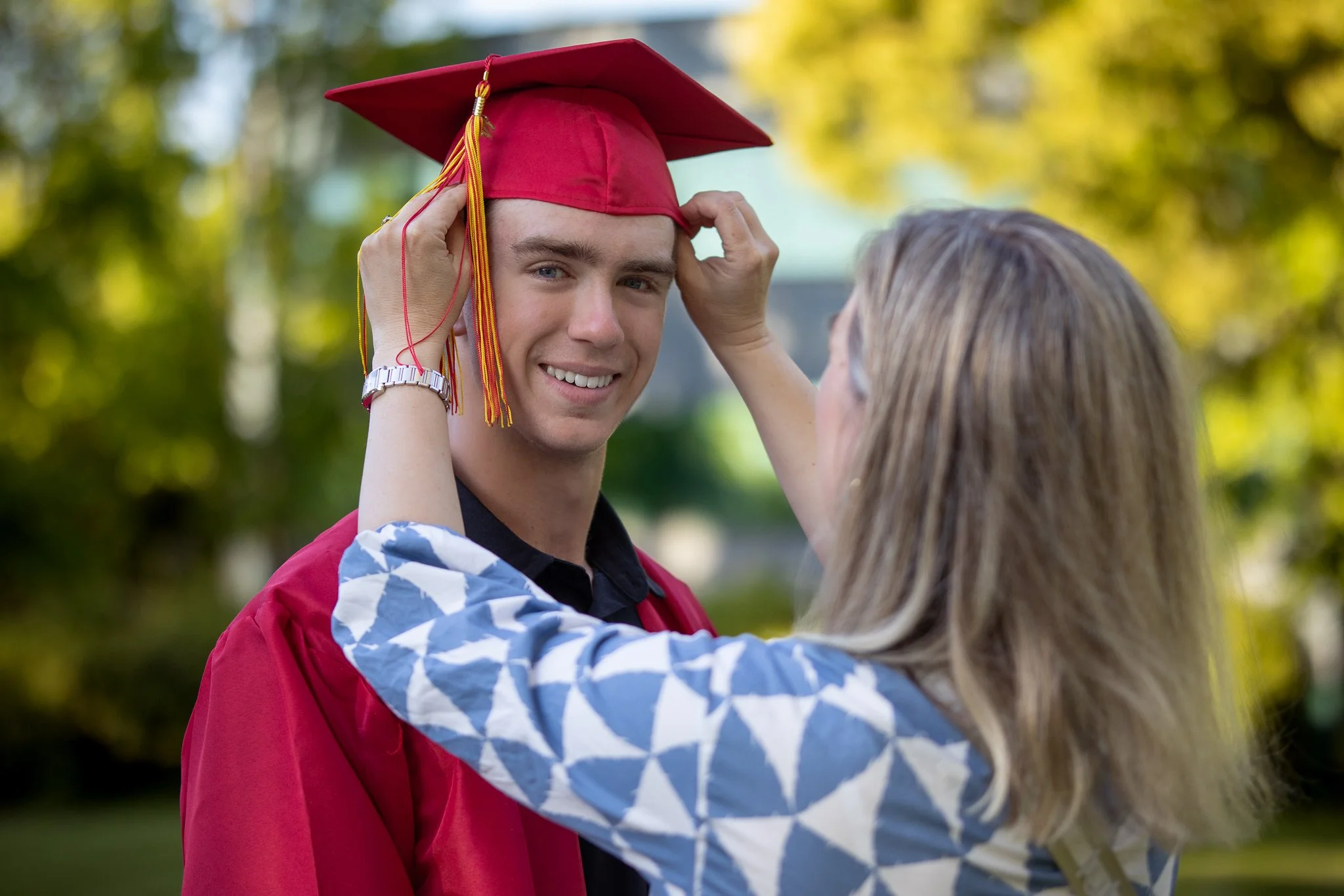 Graduate in a red cap and gown being adjusted by a woman outdoors.