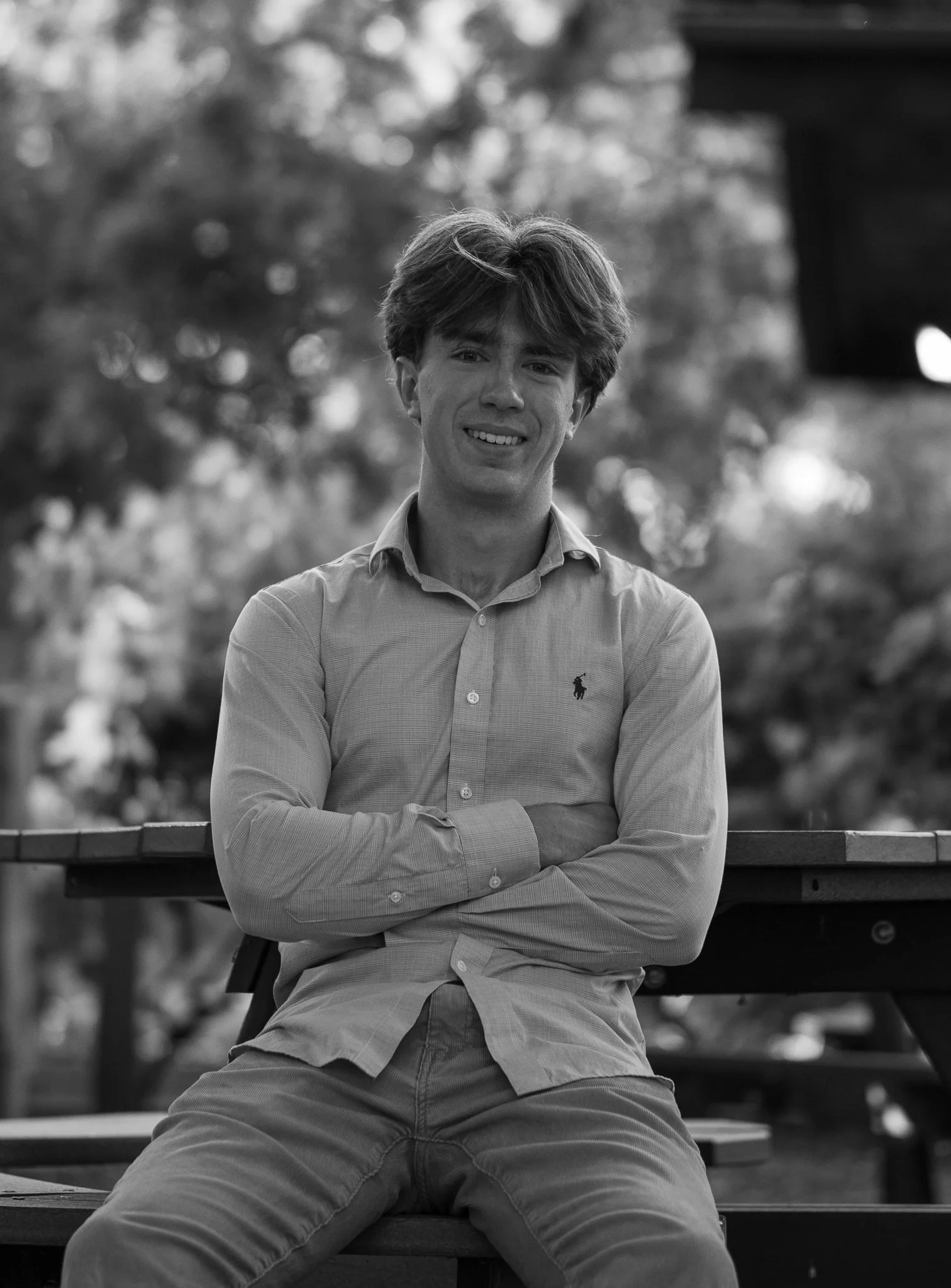 Black and white photo of a smiling man sitting at a picnic table with arms crossed, wearing a collared shirt.