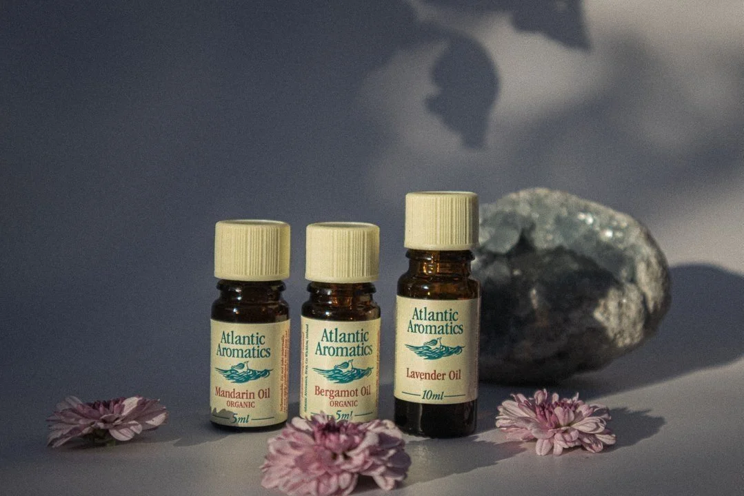 Three Atlantic Aromatics essential oil bottles (Mandarin, Bergamot, Lavender) with flowers and a stone.