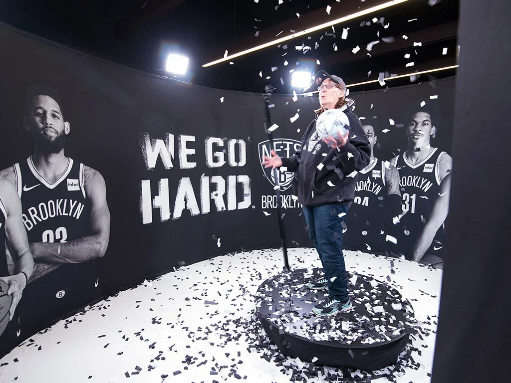 A person standing on a platform holding a basketball, surrounded by confetti, in a Brooklyn Nets-themed room with black and white photos of basketball players and the words 'WE GO HARD' on the wall.