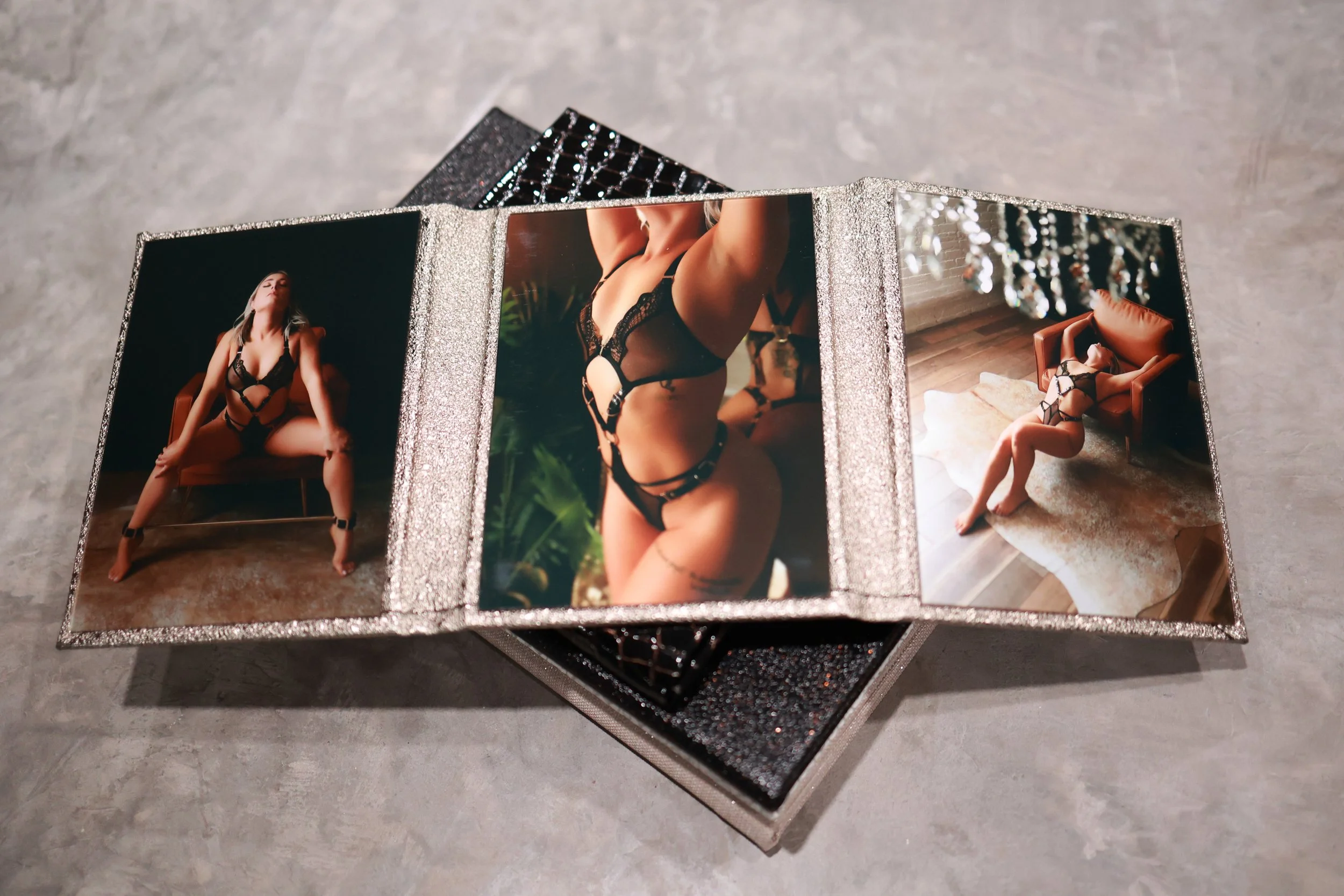 A mirrored jewelry or photo display featuring three photos of a woman in black lingerie with lace details, posed on a brown chair and in a room with ornate lighting.
